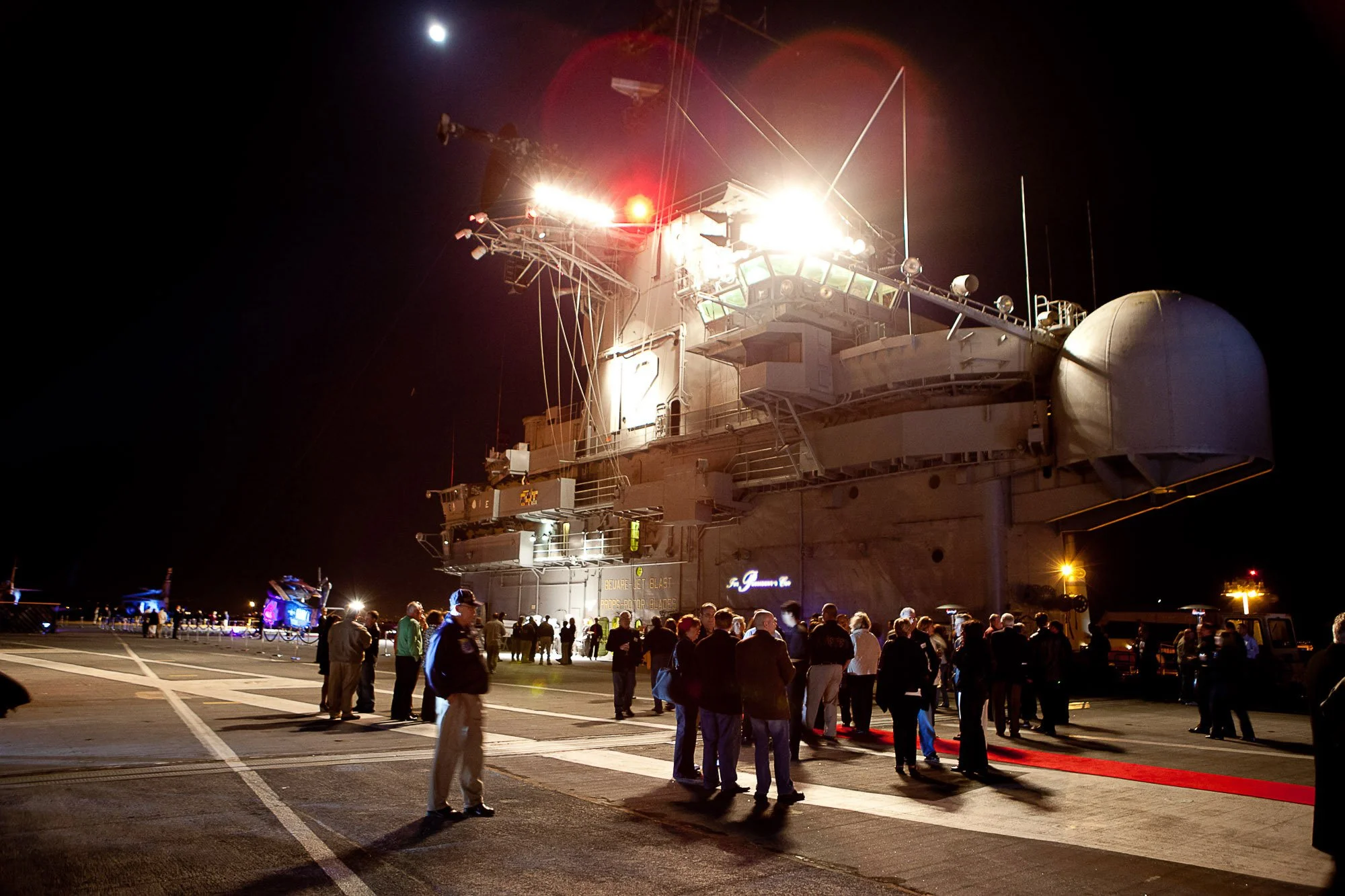 A large aircraft carrier at night, with bright lights on the deck and a group of people gathered below, some on a red carpet.