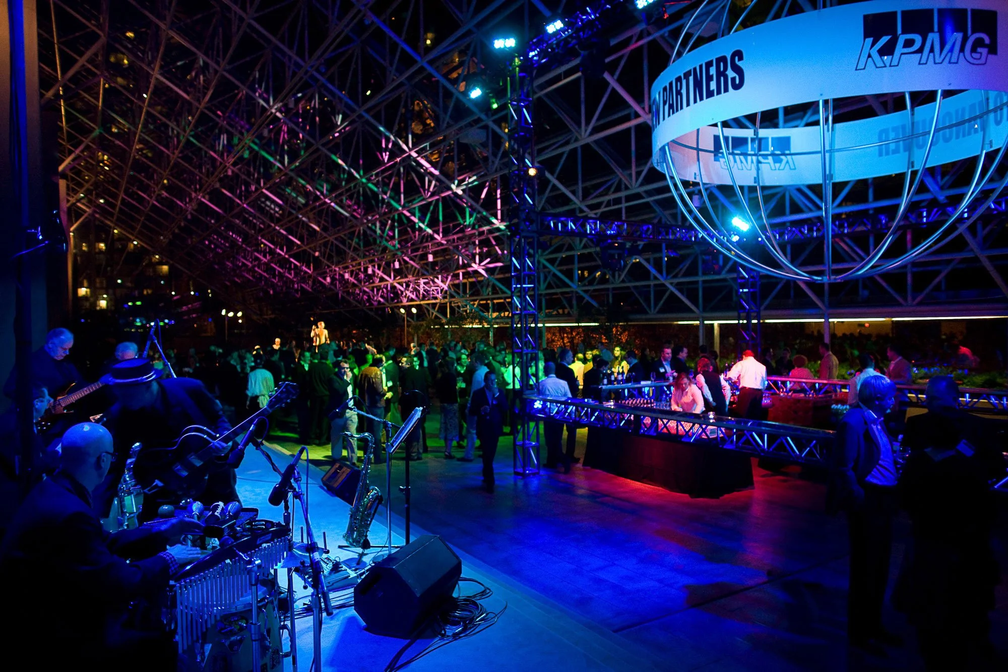 Lively indoor event with colorful lighting, a crowd of people gathered around a stage with a speaker, and a band playing instruments in the foreground.