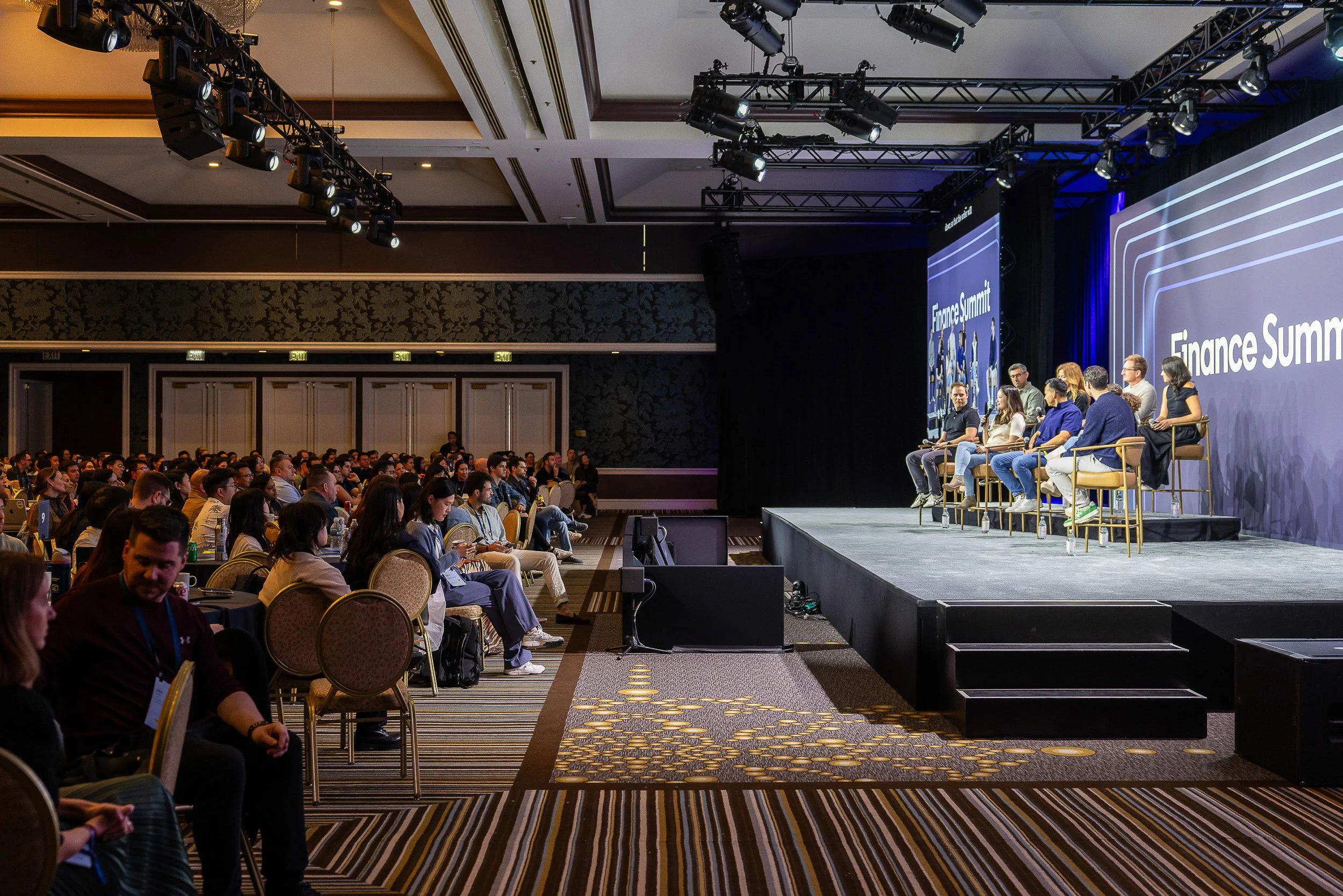 A large conference room filled with people attending a finance summit. The stage has a big screen with the text 'Finance Summit' and several panelists seated in chairs facing the audience.