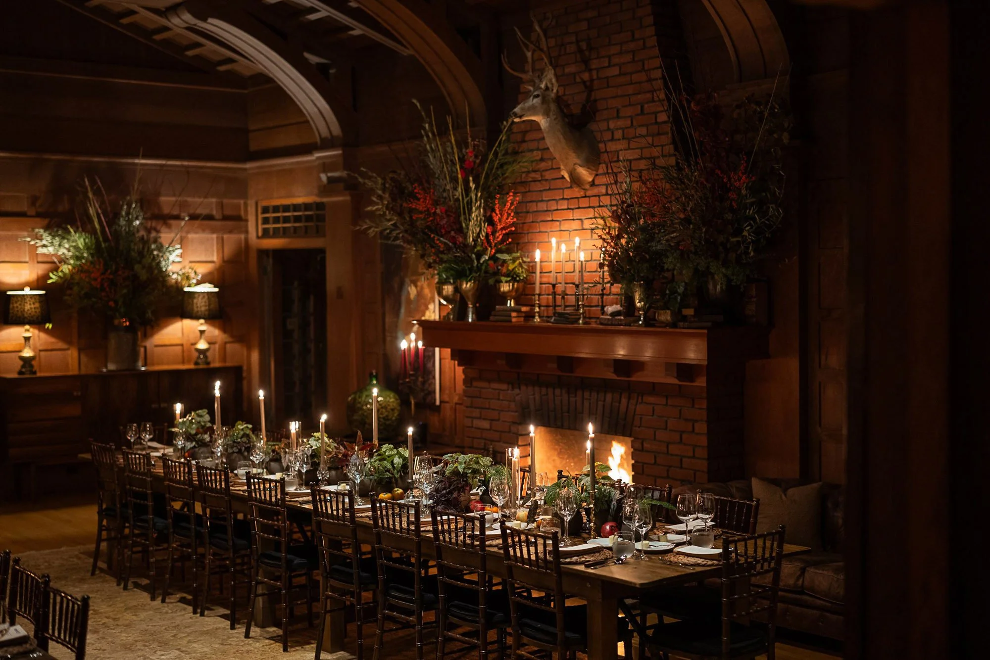 A long dining table set for a formal dinner with candles and greenery in a cozy wood-paneled room with a brick fireplace, decorated for a festive occasion.