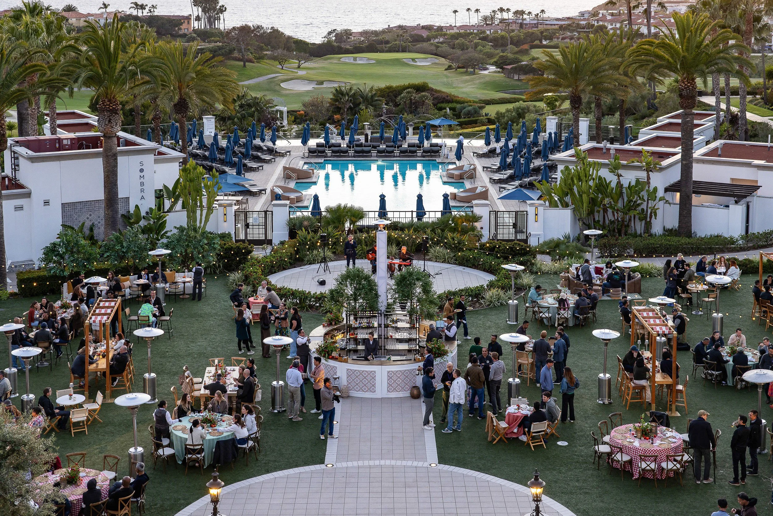 People enjoying an outdoor event at a restaurant with a pool, surrounded by palm trees and overlooking a golf course near the water.