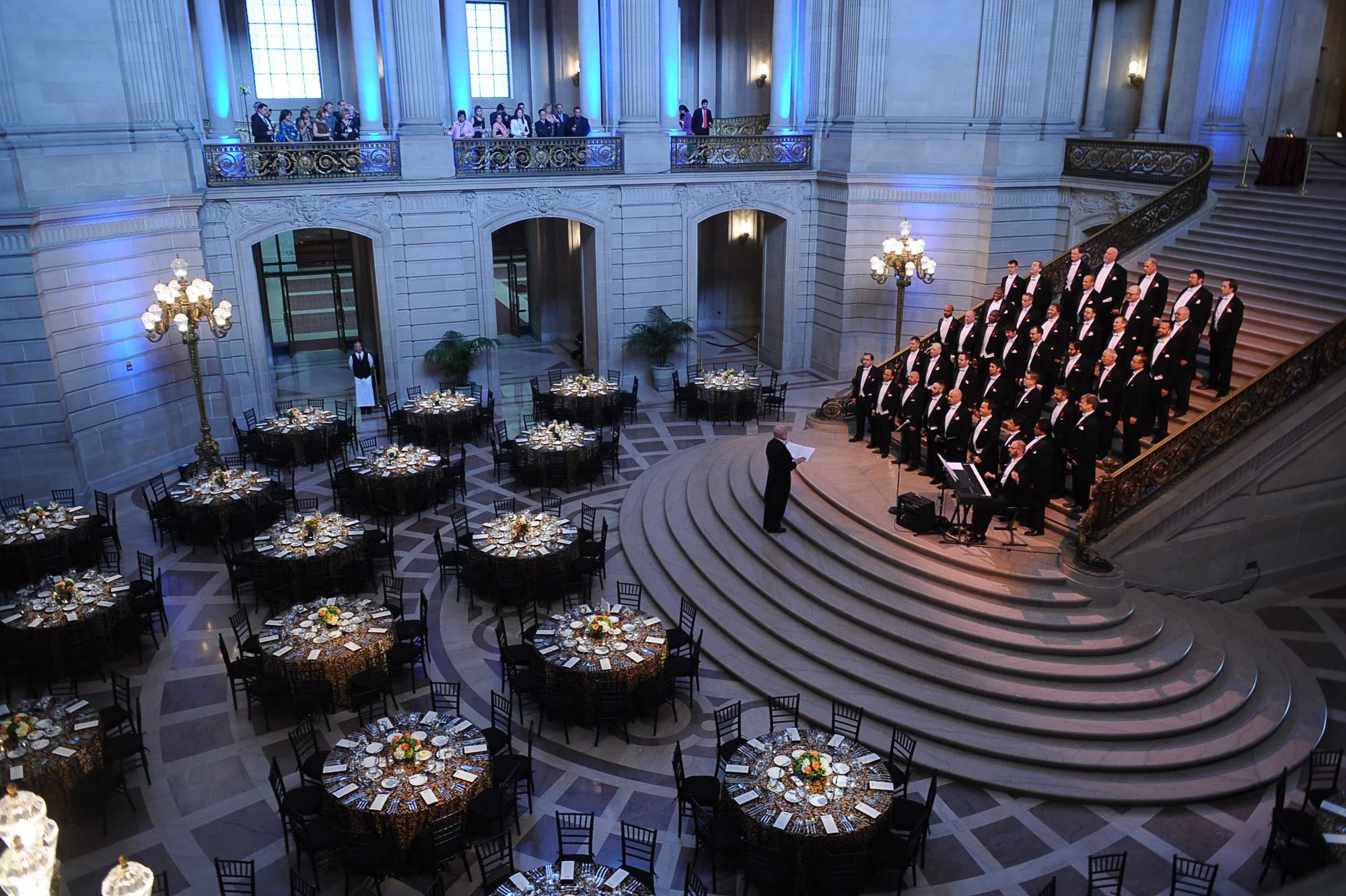 A large, elegant hall with high ceilings, tall windows, and ornate architectural details. There are multiple round banquet tables decorated with floral centerpieces, set with plates and utensils. A choir dressed in black tuxedos with white shirts is 