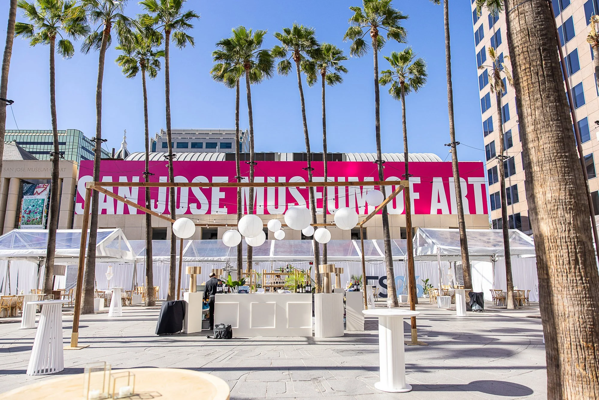 Outdoor event space with white tents, decorative paper lanterns, and tall palm trees in front of a large pink sign that reads 'San Jose Museum of Art.' Modern buildings are visible in the background under a clear blue sky.
