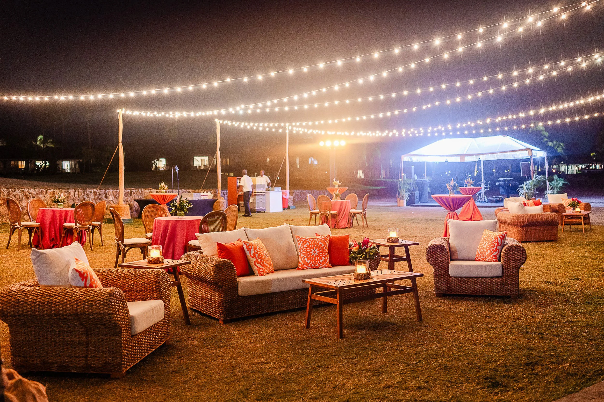 Outdoor nighttime event with string lights overhead, tables with red tablecloths, and cozy seating with orange and white cushions.