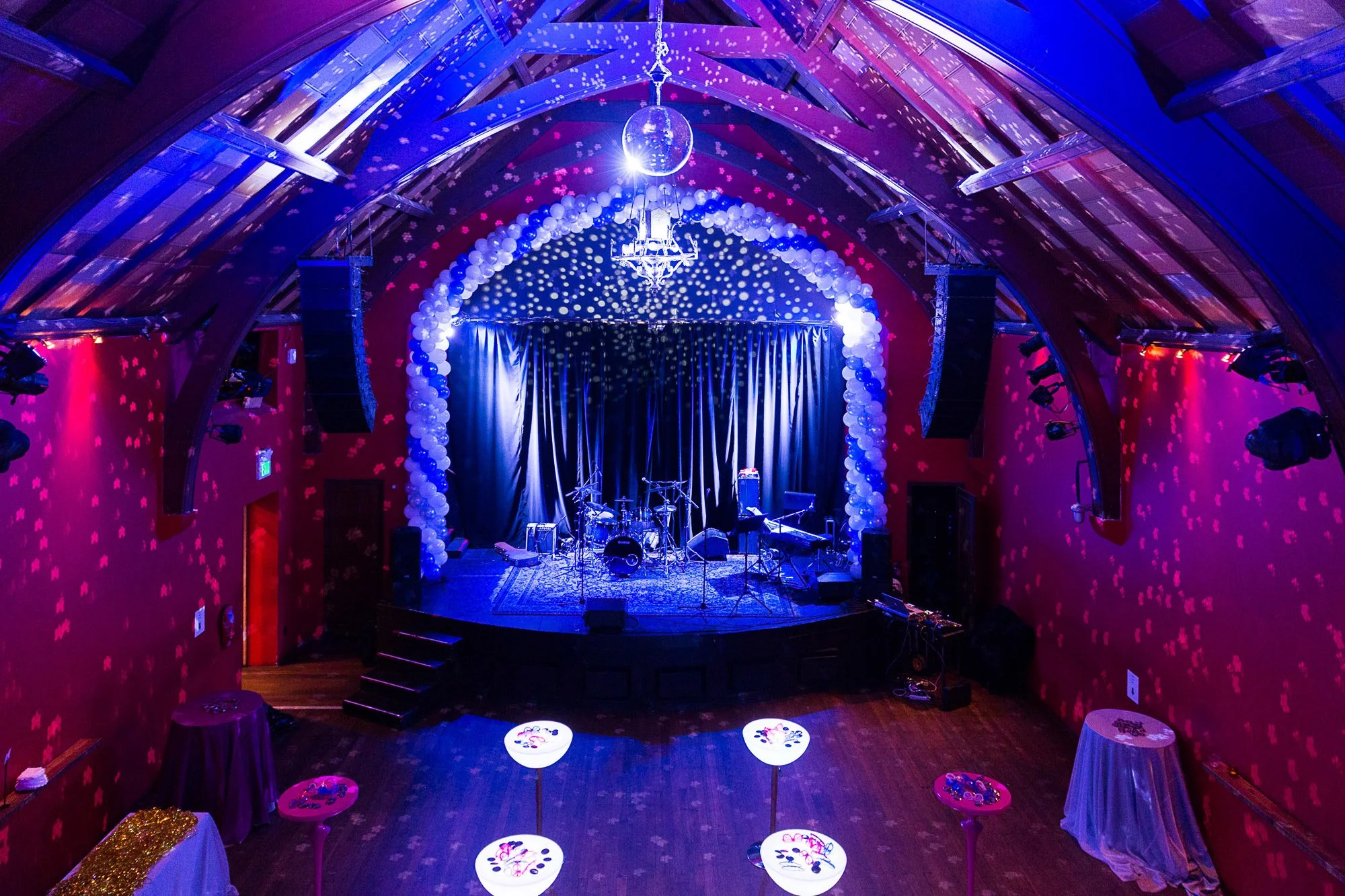 Concert stage decorated with blue and white balloons, a glittering disco ball hanging from the ceiling, and purple and pink lighting. The stage is set with musical instruments, including drums, keyboards, and microphones, with small tables and tables