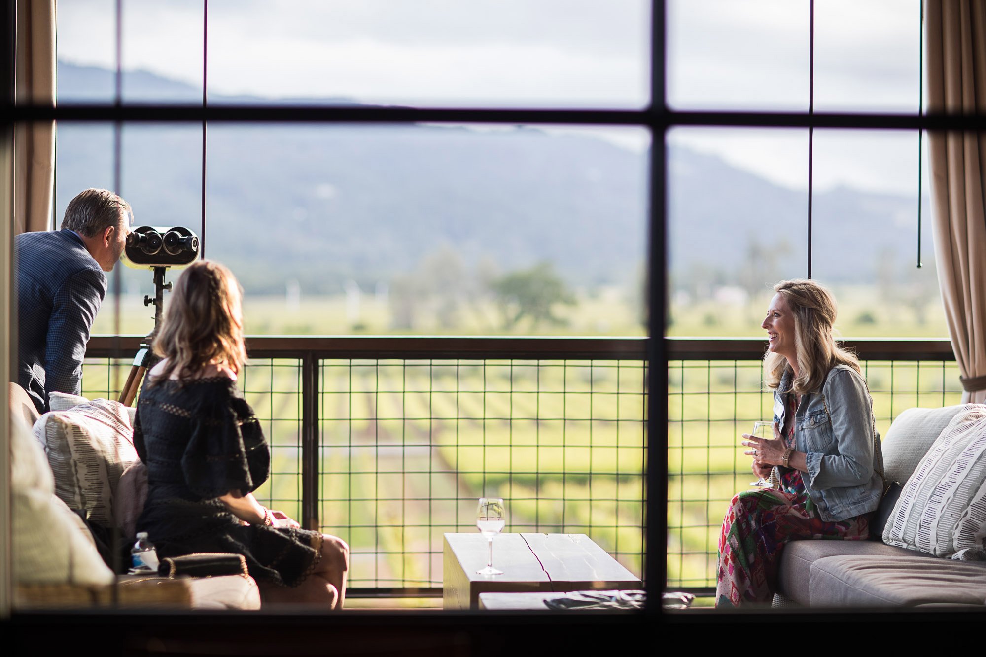 People engaging in a conversation on a balcony near large windows, overlooking a green landscape and mountains.