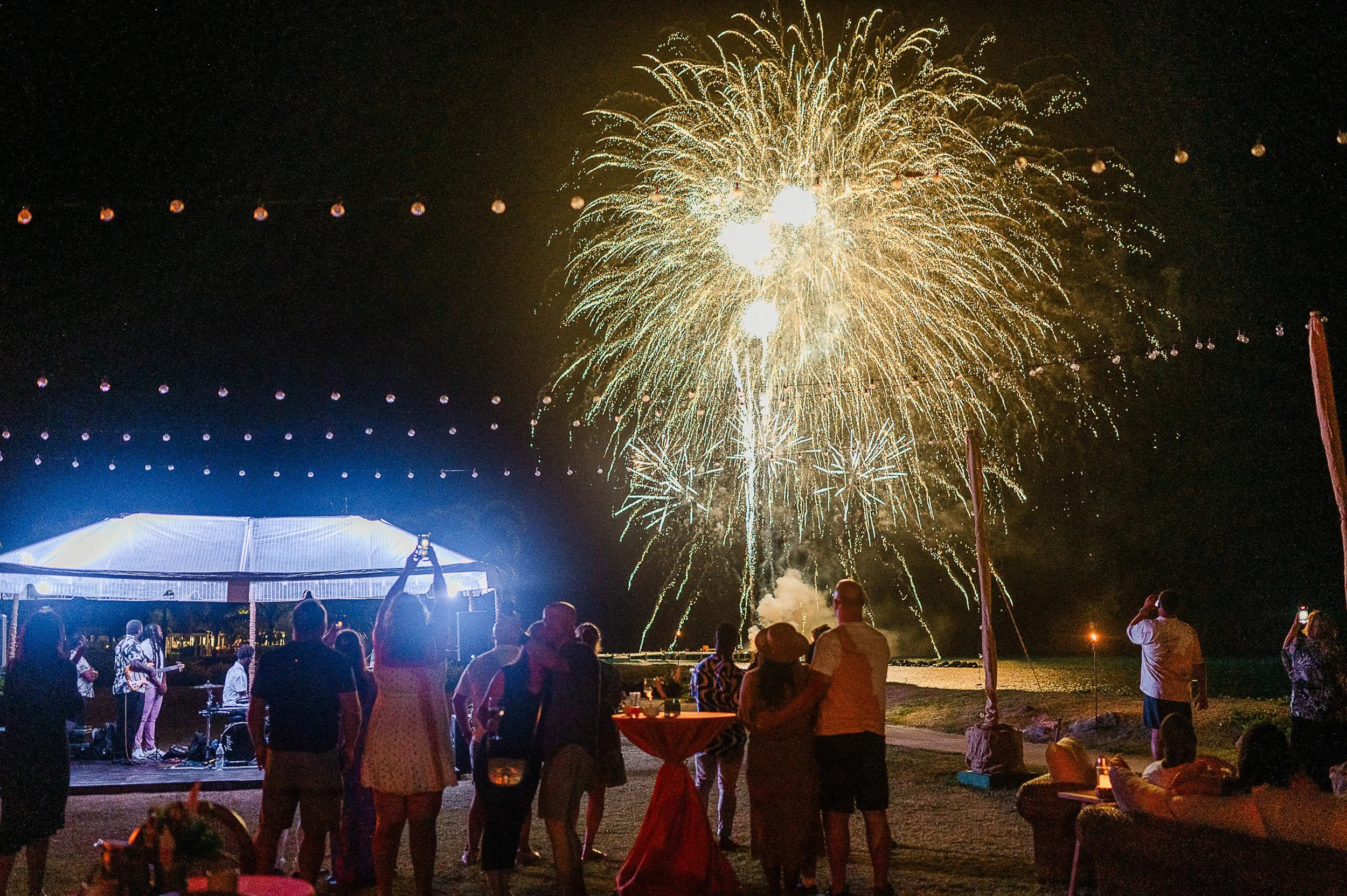 People watching fireworks display at night, with illuminated tents and string lights overhead.