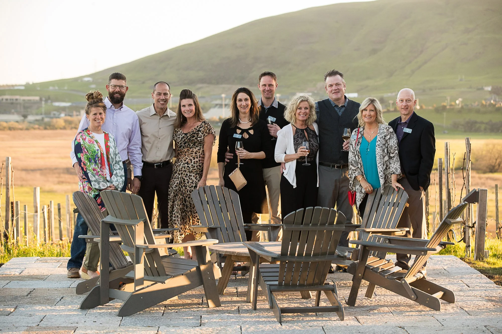 Group of ten people standing outdoors with chairs, holding glasses of wine, against a scenic hilly landscape during sunset.