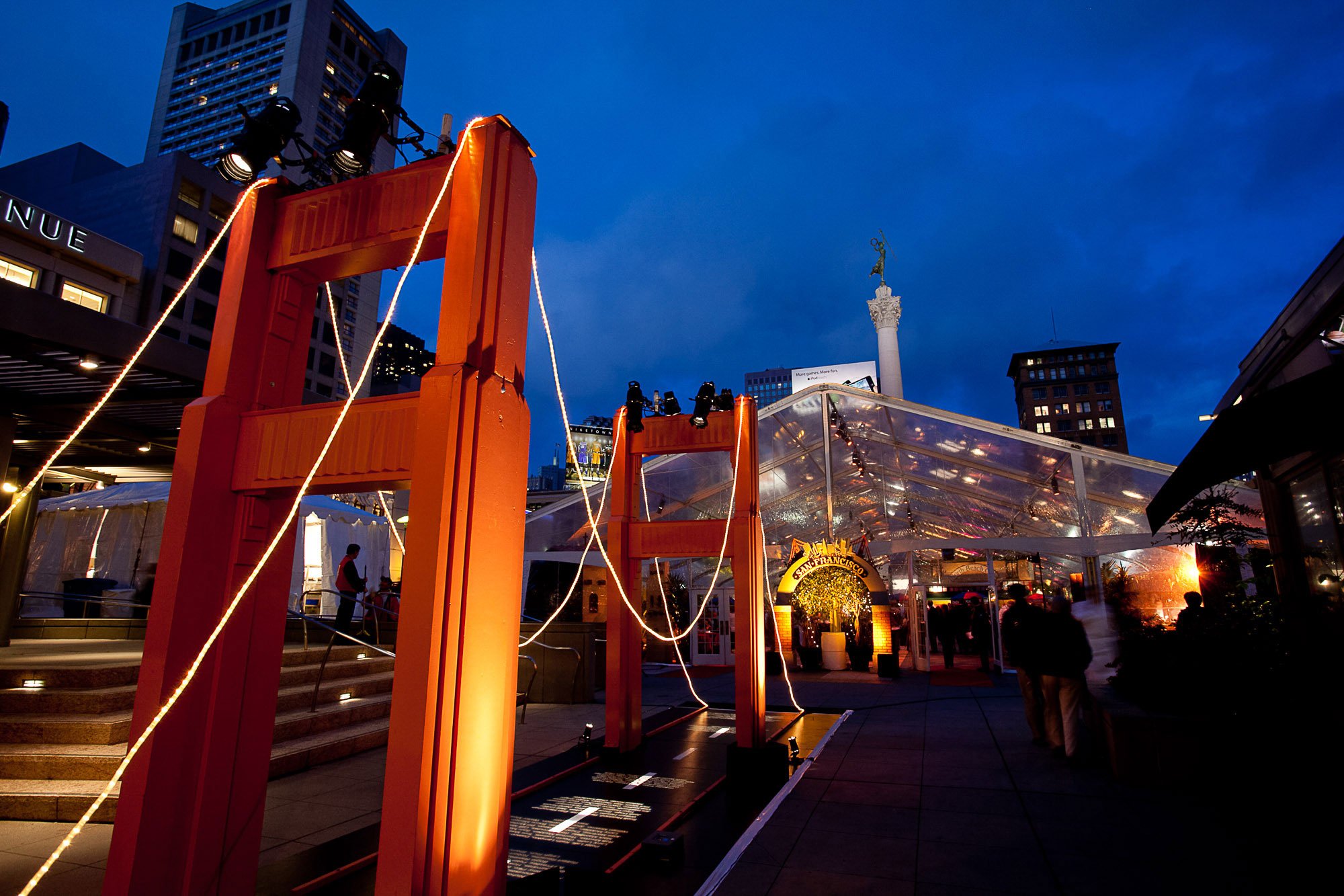 An outdoor city scene at dusk featuring a large red structure, likely a scale model of the Golden Gate Bridge, with string lights. In the background are tall buildings, a statue atop a column, a glass canopy, and a sign that says 'San Francisco' with
