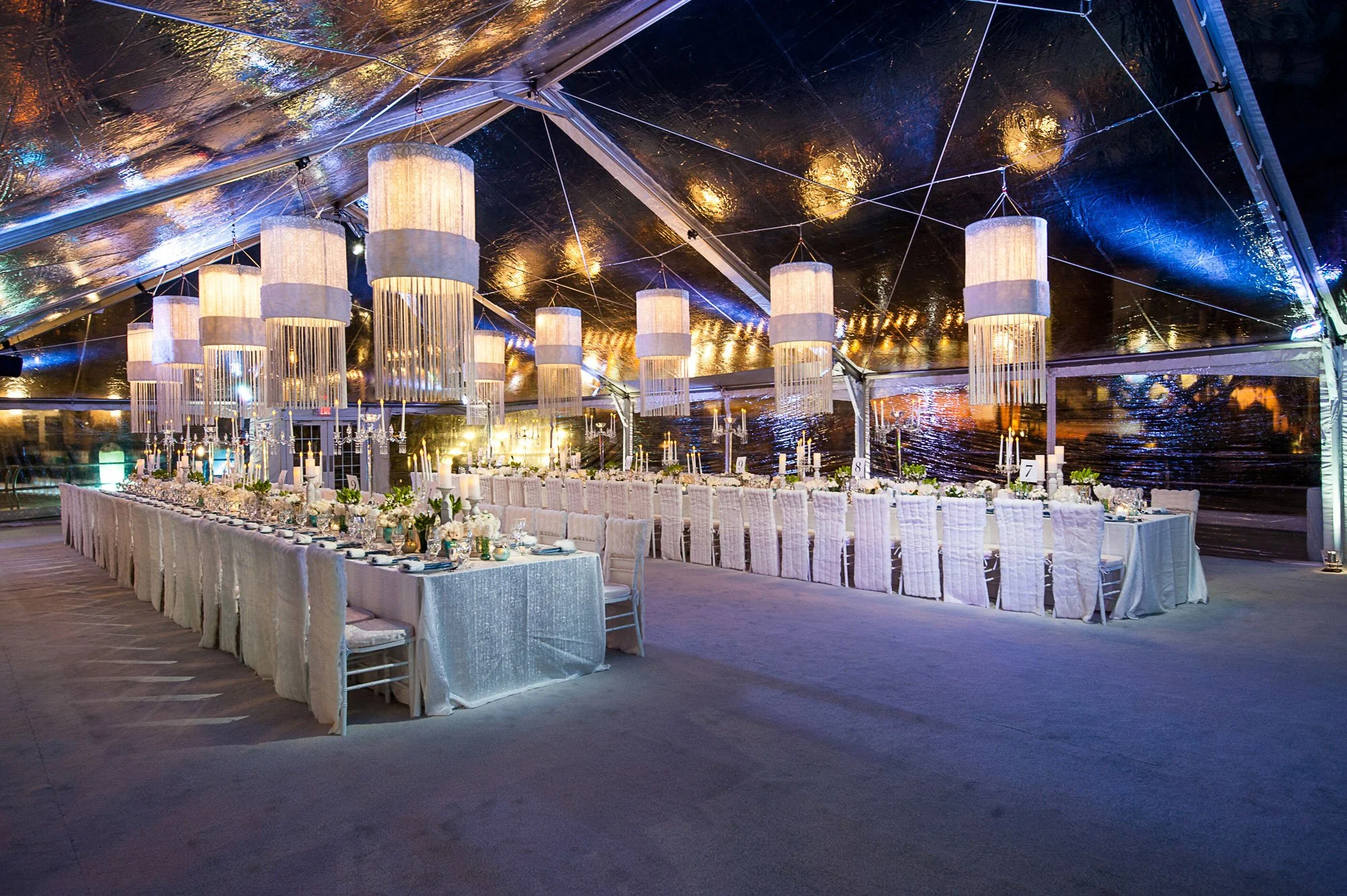 Elegant banquet table setup inside a glass tent with hanging pendant lights, candles, and floral centerpieces, overlooking a body of water at night.