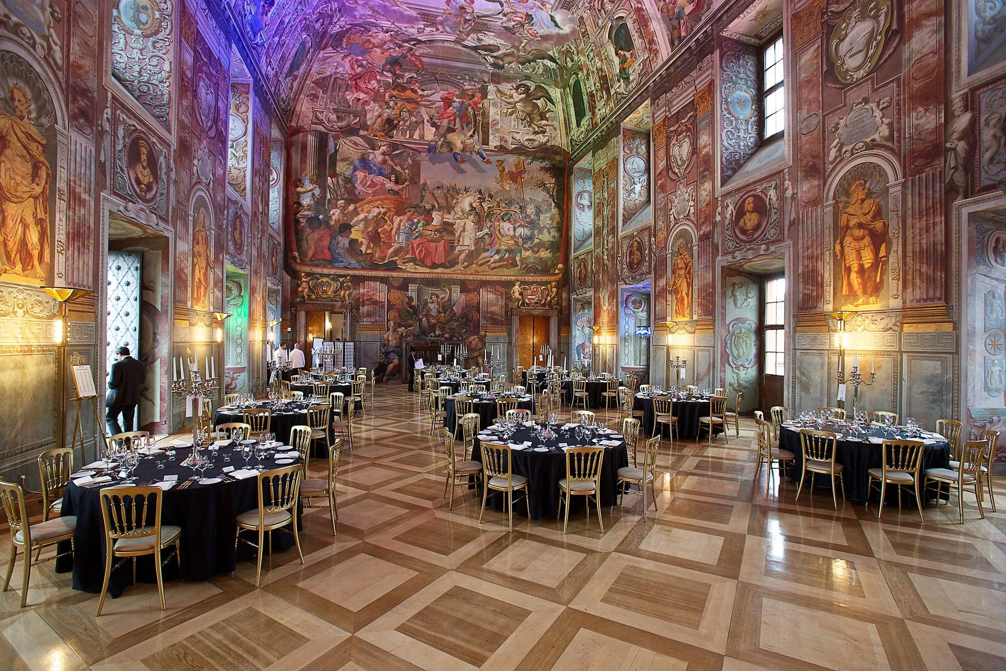 Elegant banquet hall with tall ceilings, ornate frescoes and murals, large windows, and round tables covered with black tablecloths, set for a formal event.