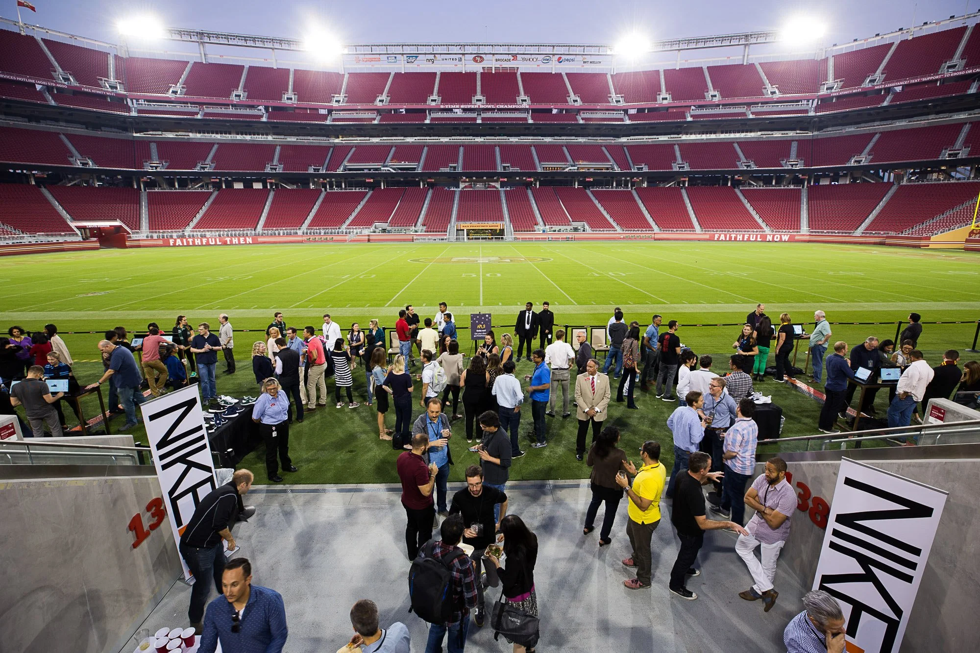 People gathering inside a football stadium, view from the upper level looking down onto the field, which is surrounded by a seating area with red seats. The field is empty, with some groups of people standing and talking near the field and concession