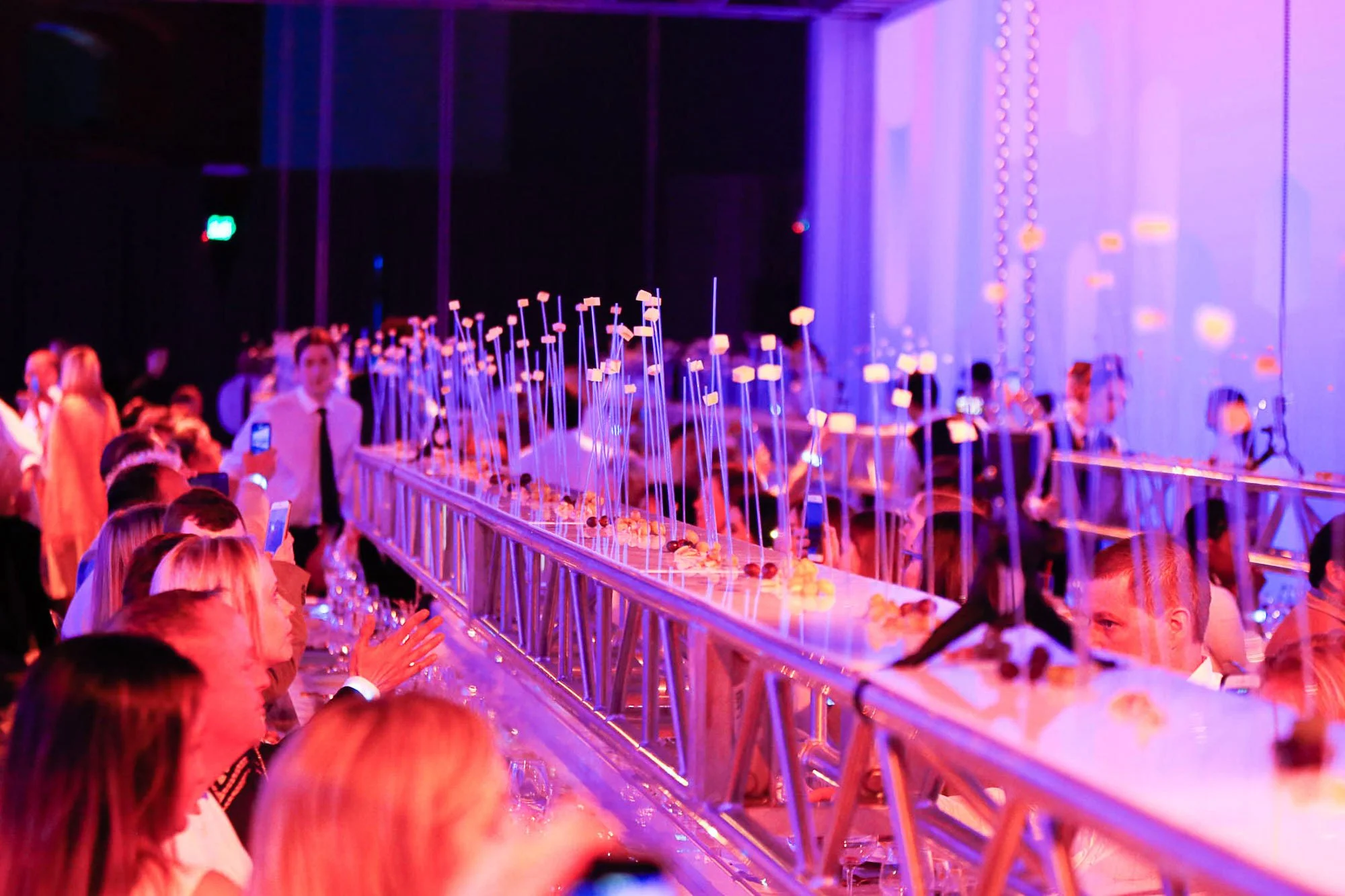A crowded indoor event with purple lighting, showcasing a long table with desserts arranged on tall sticks, where people are seated and some are taking photos.