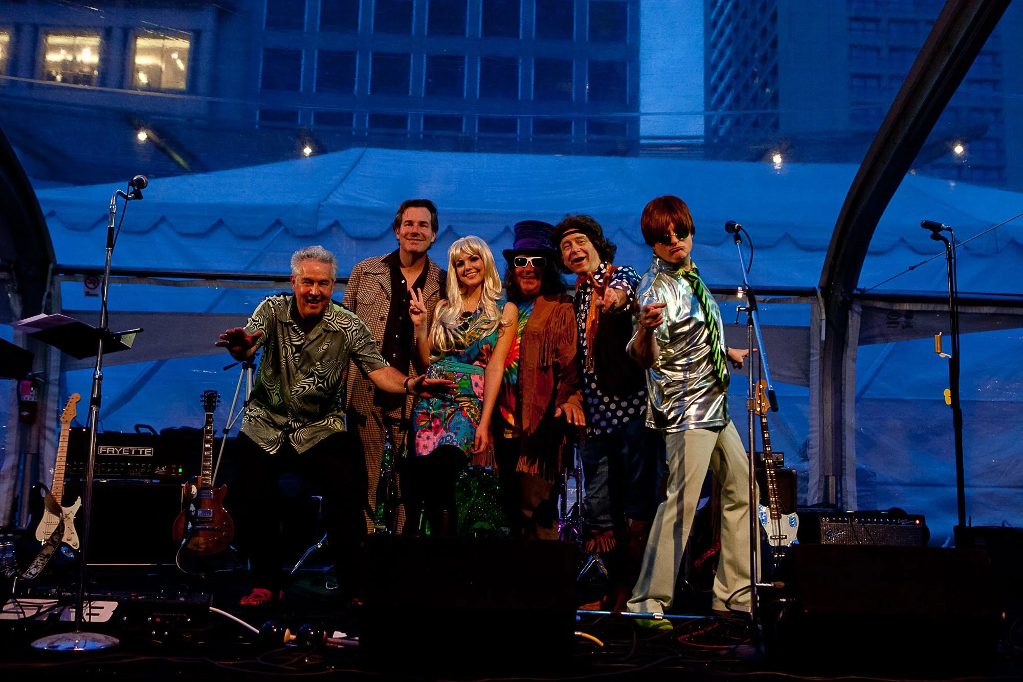 A group of seven performers dressed in colorful and eccentric outfits on a stage, posing for a photo at an outdoor music event at night, with musical instruments and equipment visible around them.