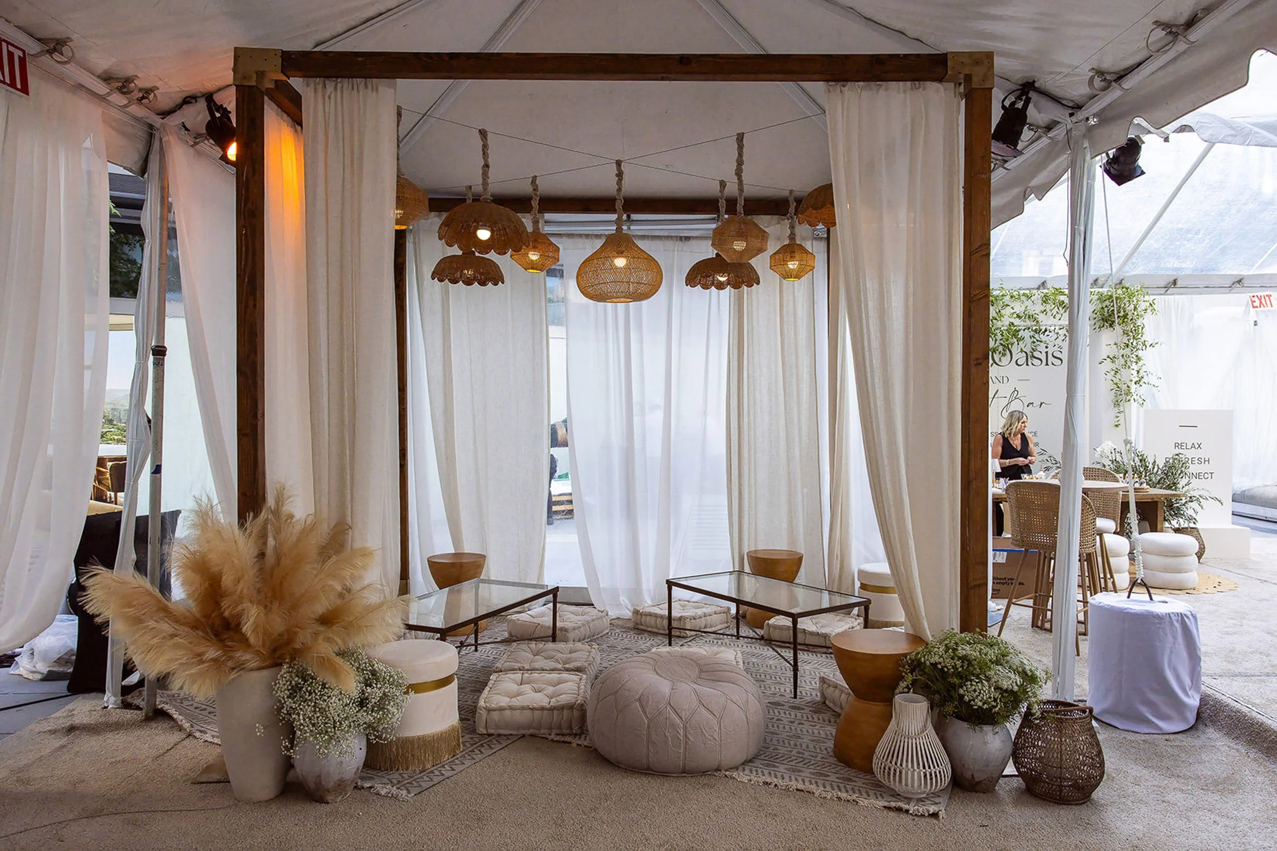 A cozy seating area with wicker lamps hanging from the ceiling, surrounded by white curtains, patterned rugs, and decorated with potted plants and plush cushions.
