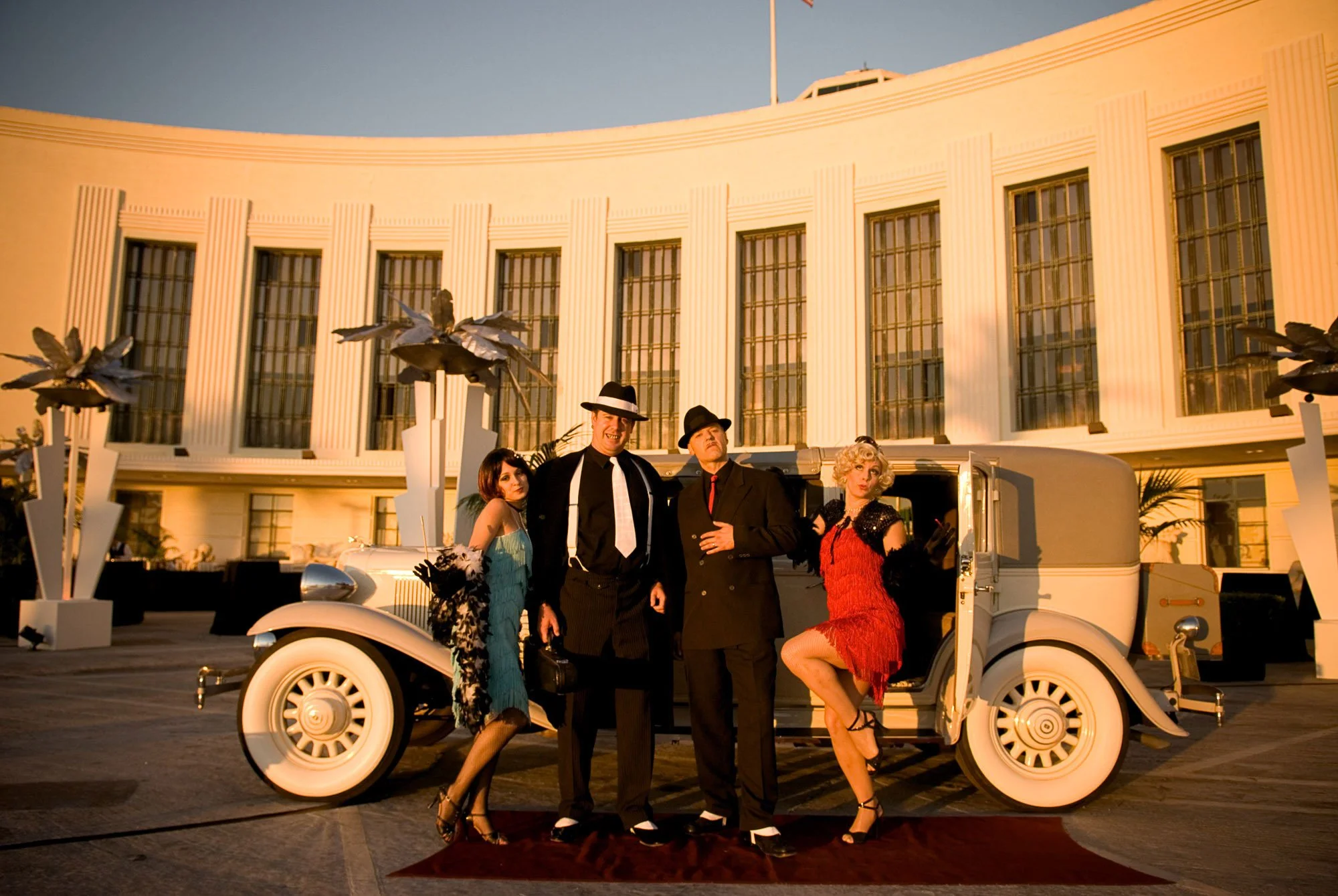 Four people dressed in 1920s style clothing posing on a vintage white car in front of a art deco building with large windows and flowers sculptures.
