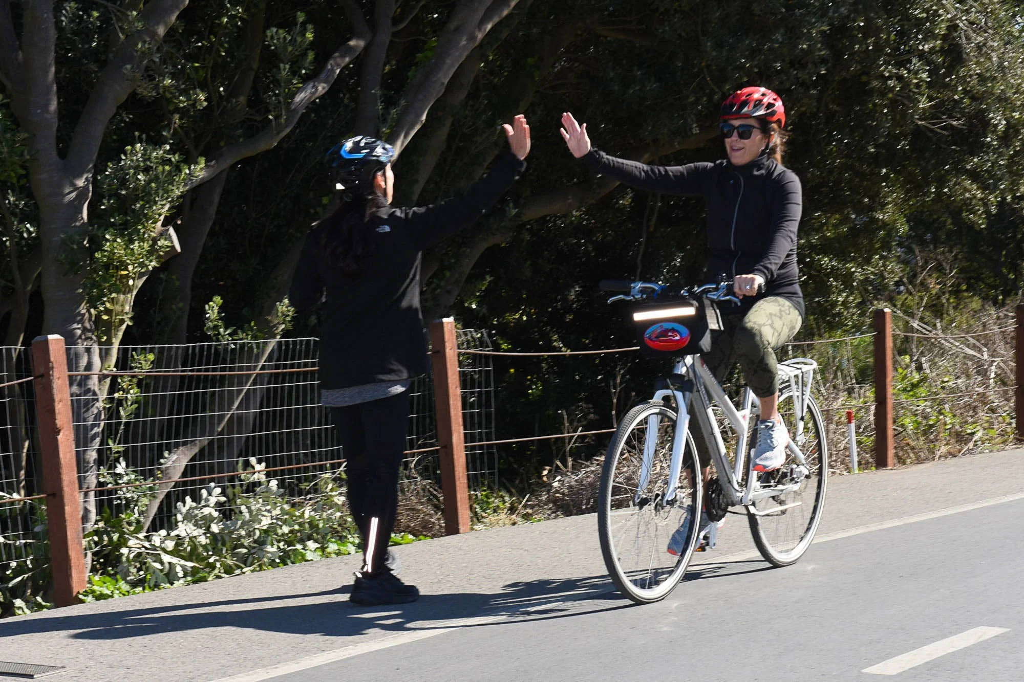 Two women high-five each other while one rides a bicycle on a paved path next to a wooden fence and trees.