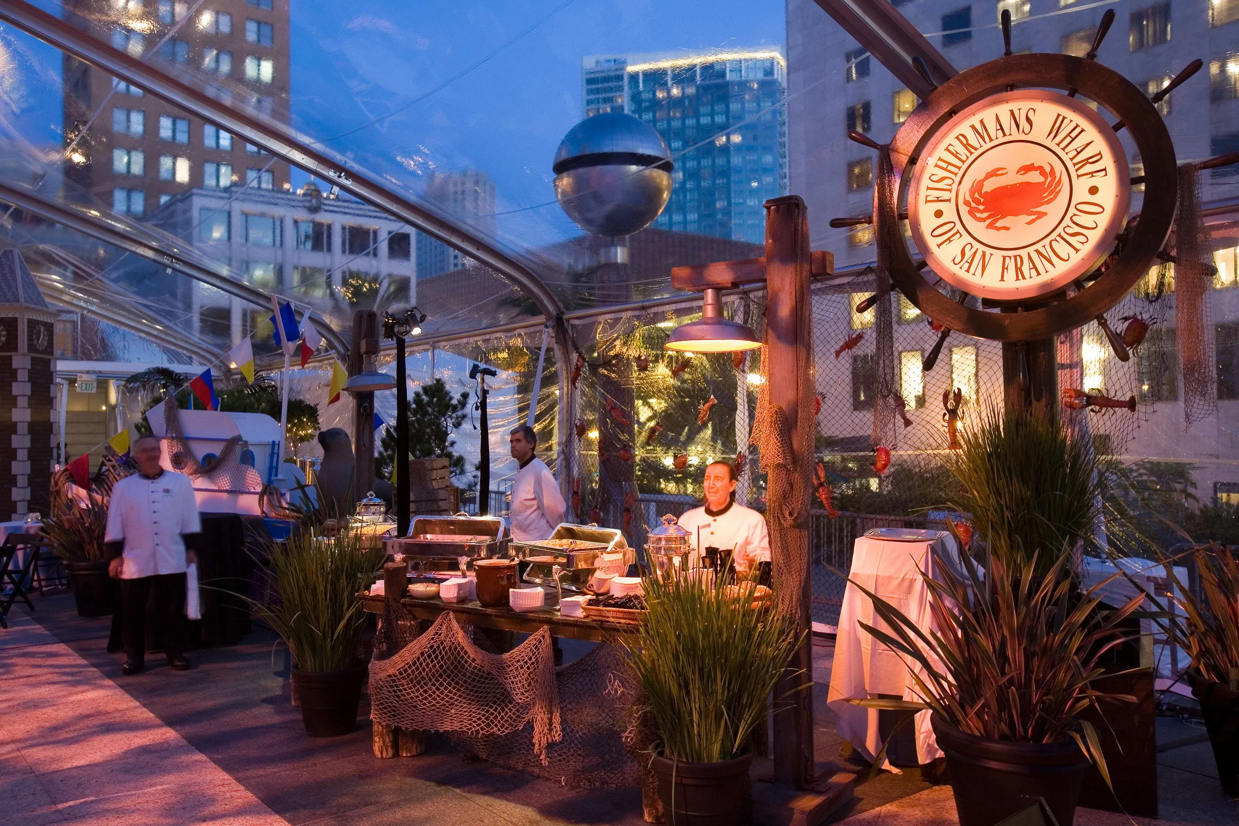 Outdoor restaurant or event space with chefs and food stations, decorated with potted plants, under a tent with city buildings in the background, during evening or dusk.
