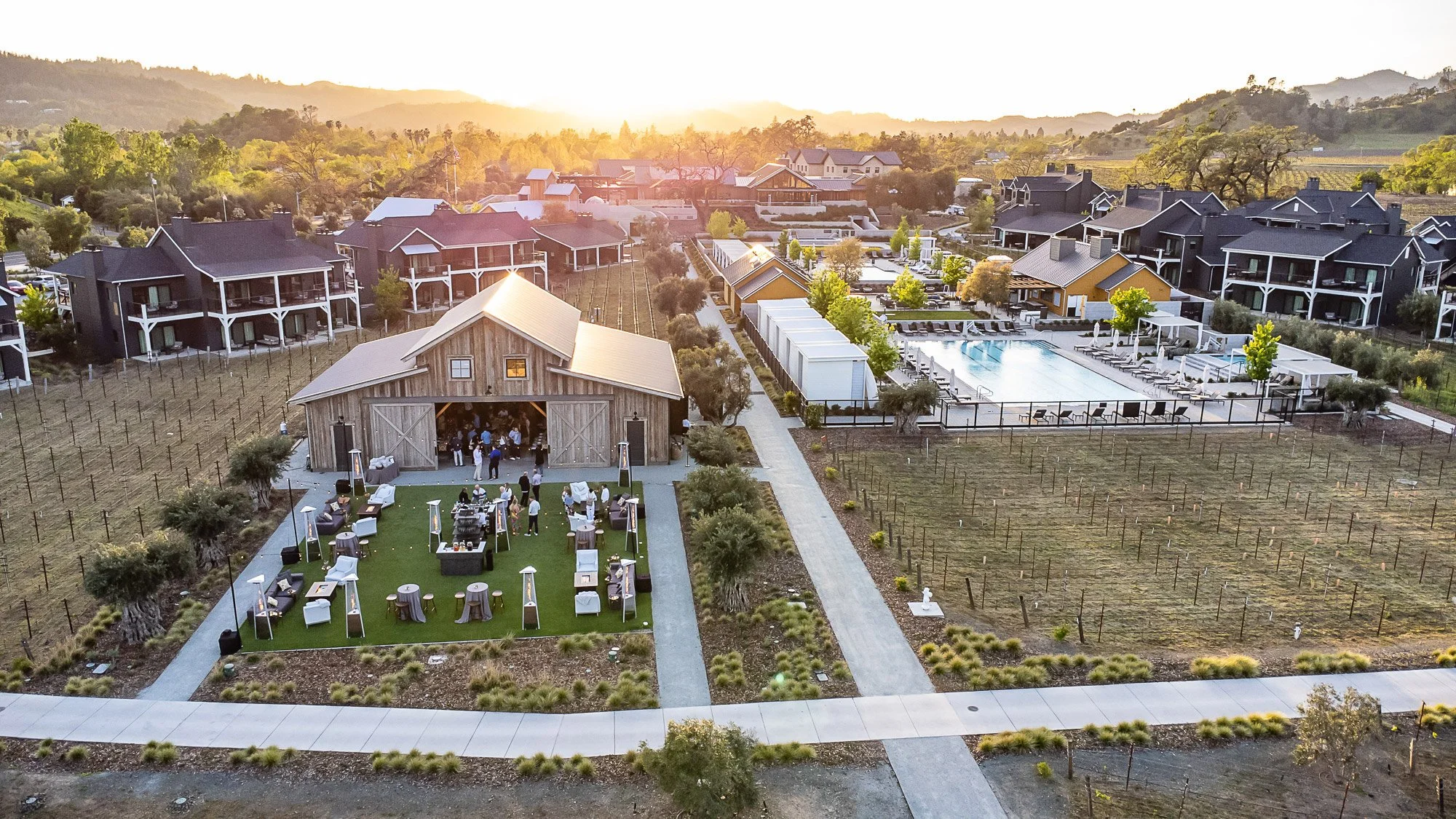 An aerial view of an outdoor event at a winery or vineyard, with a rustic barn, patio area with seating, a large swimming pool, and residential-style buildings in the background during sunset.