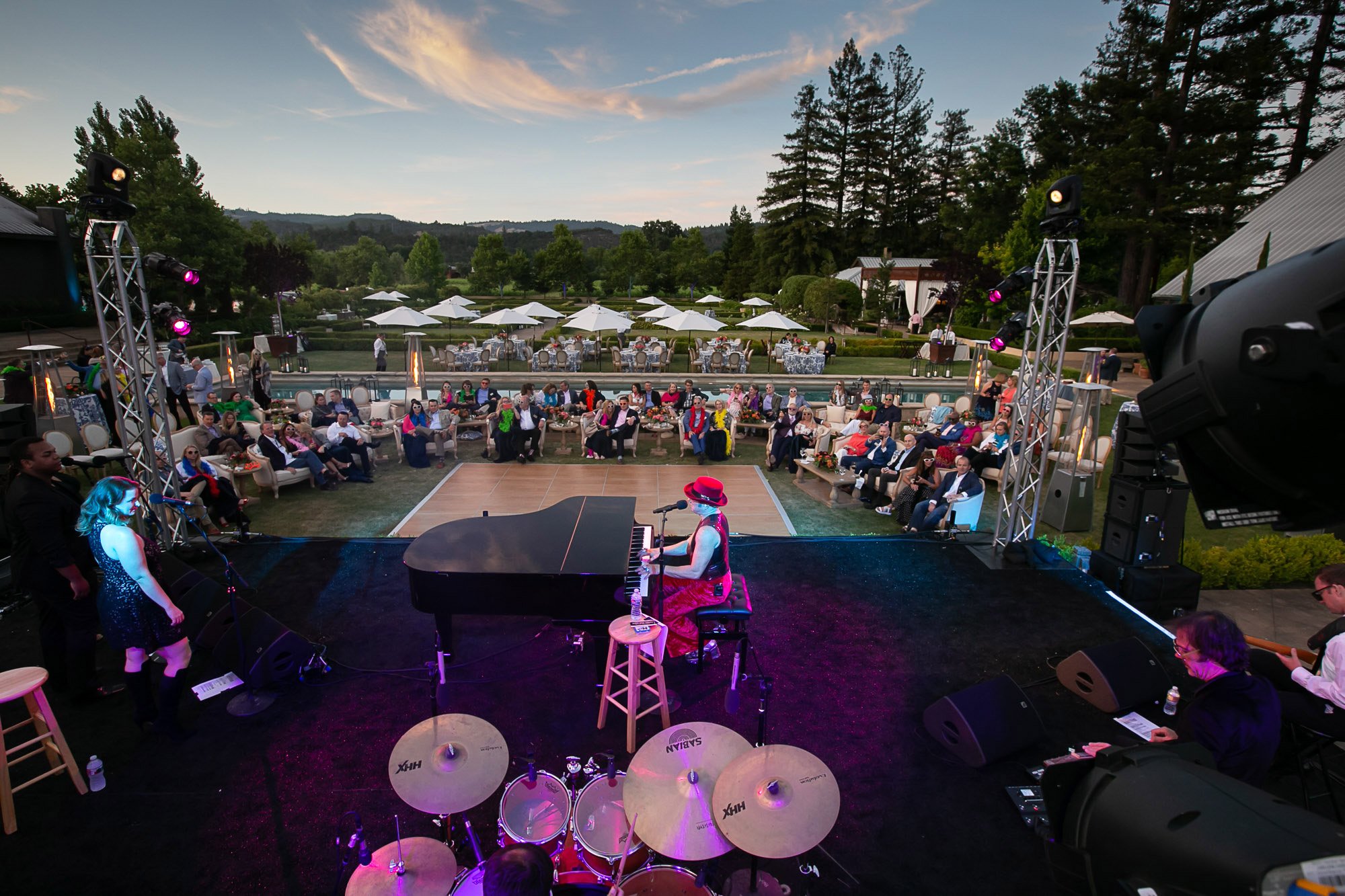 An outdoor live music concert with a pianist and singer on stage, audience seated on couches and chairs, and a scenic background featuring trees, a pond, and a sunset sky.