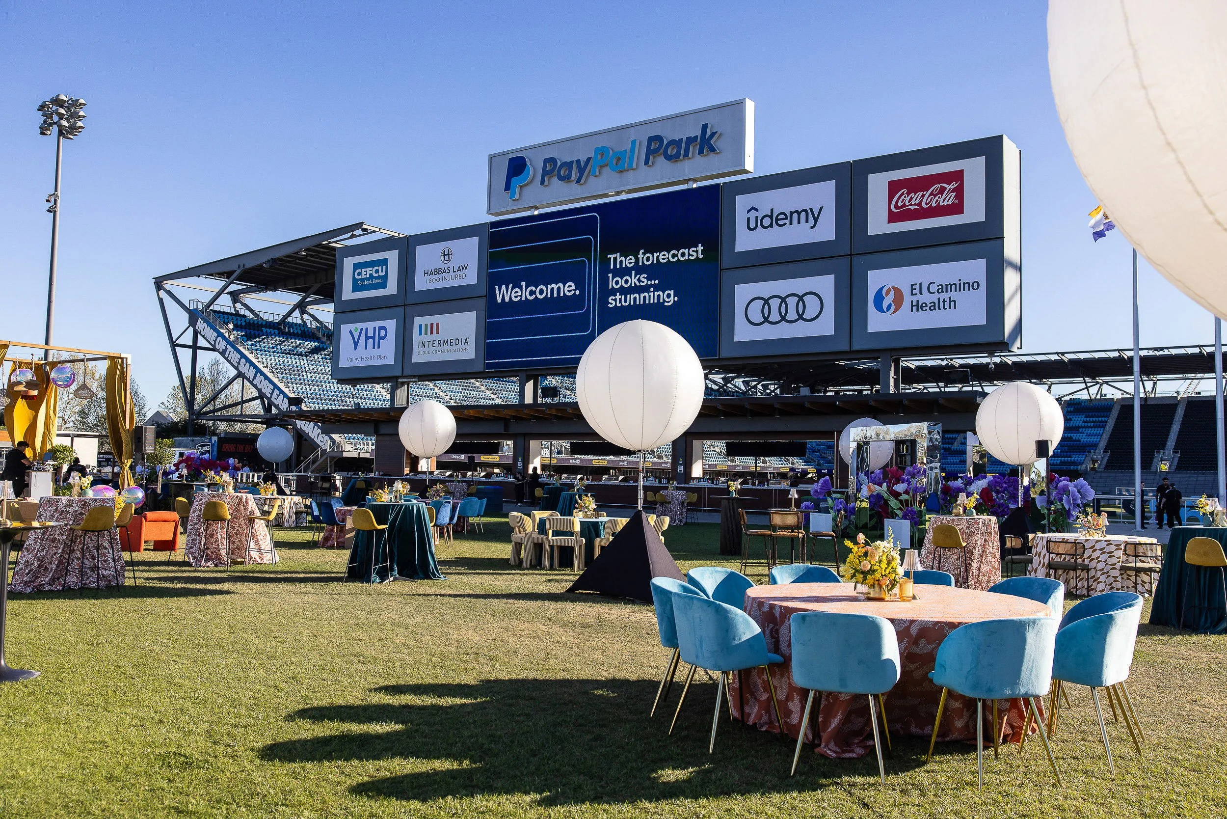 Outdoor event space at PayPal Park, with colorful tables and chairs, large paper lanterns, blooming flowers, a large digital scoreboard with advertisements, and a clear blue sky.