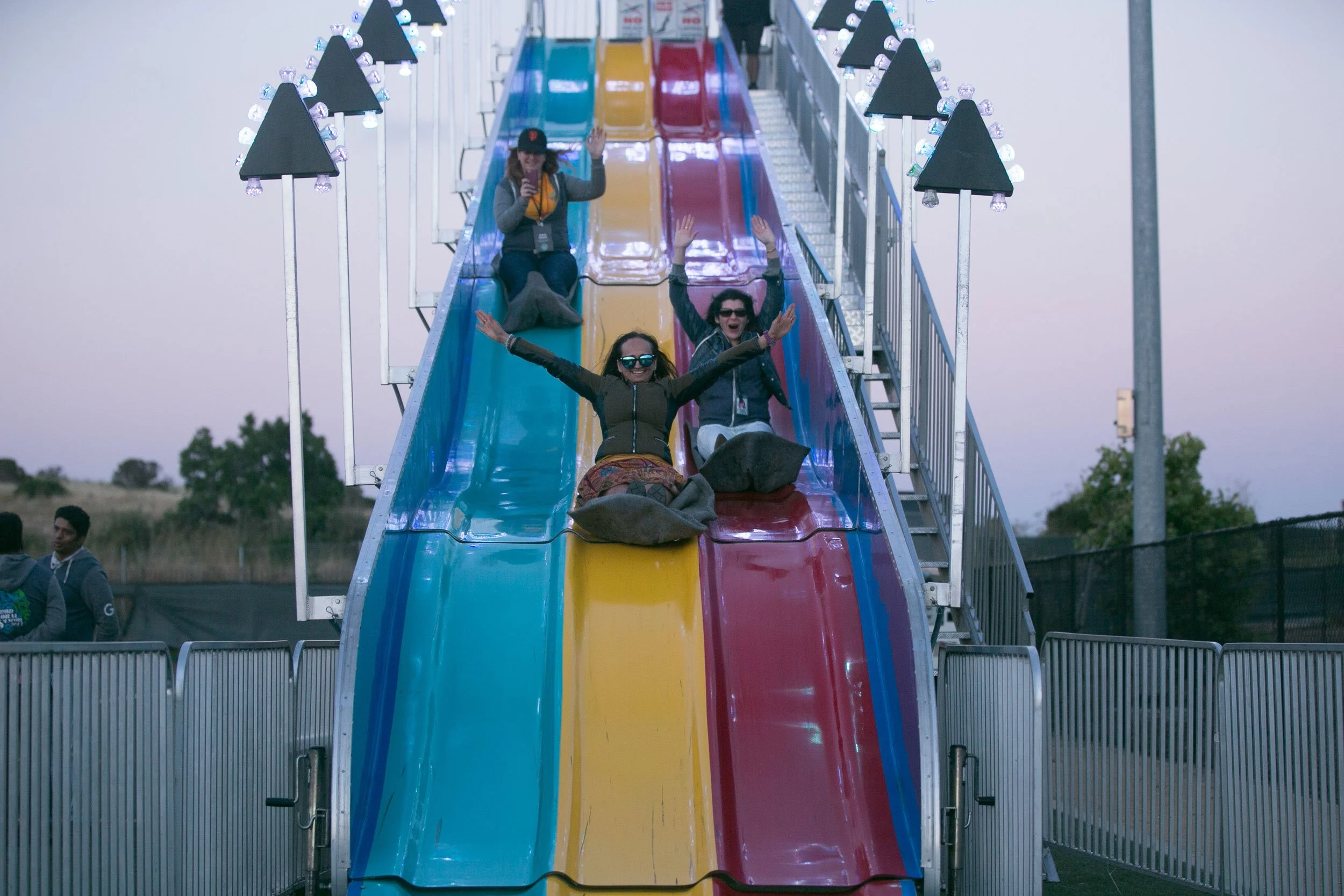 People enjoying themselves on a colorful amusement park slide with three lanes in yellow, blue, and red, surrounded by decorative black triangular signs with lights, during the daytime.