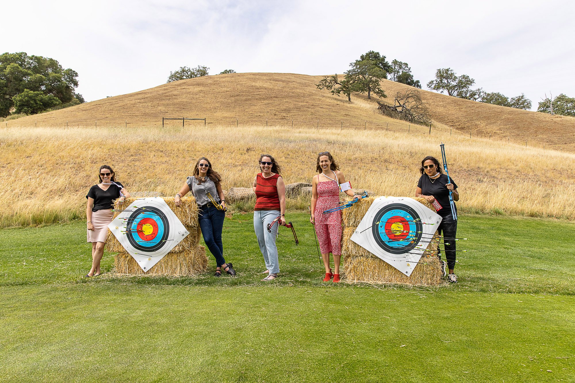Four women standing in front of hay bales with archery target banners, holding archery equipment, outdoors in a grassy area with rolling hills and trees.