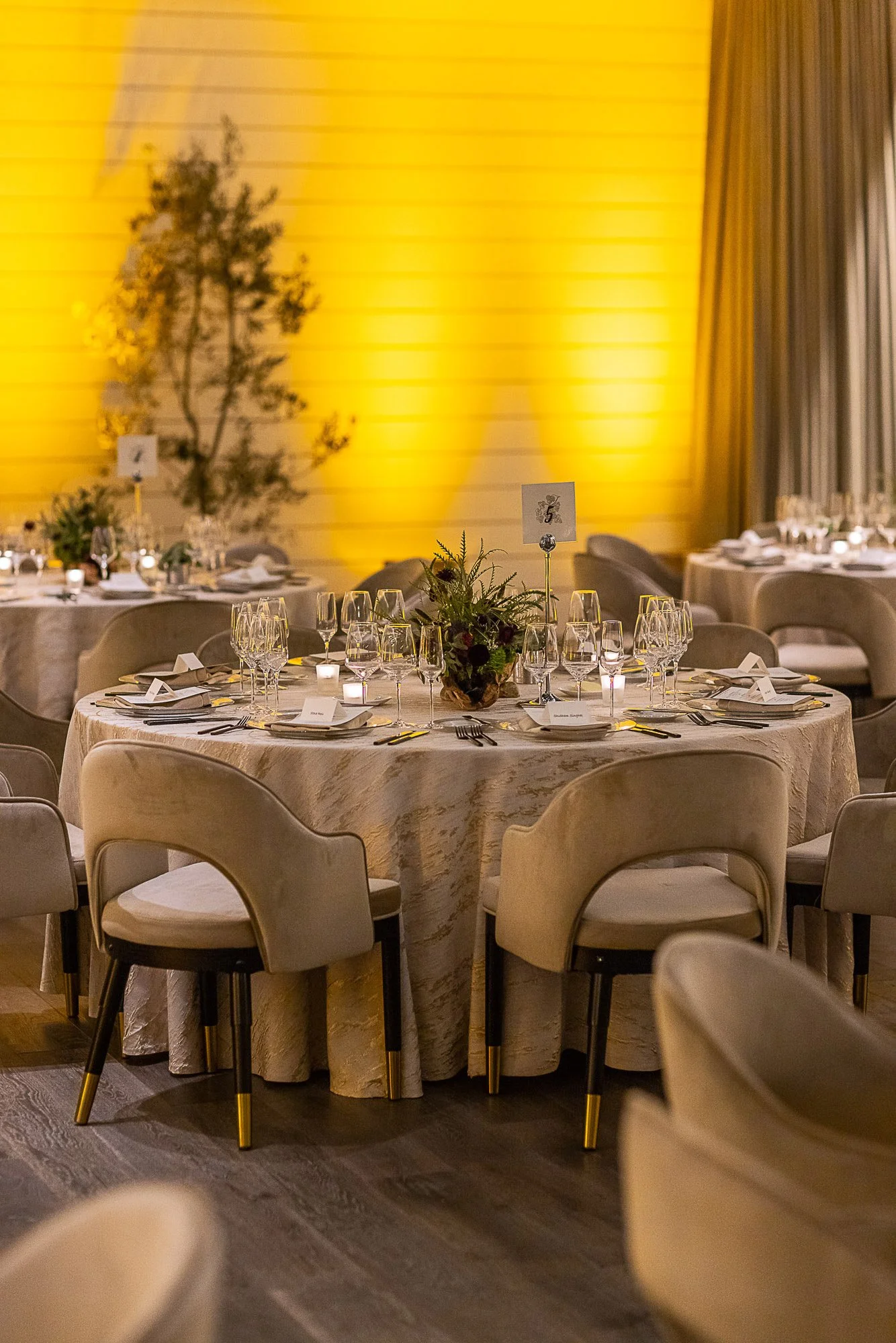 Formal dining setup with a round table, beige chairs, white tablecloth, wine glasses, candles, and floral centerpiece. The background features a yellow wall with a shadow of a tree and beige curtains.
