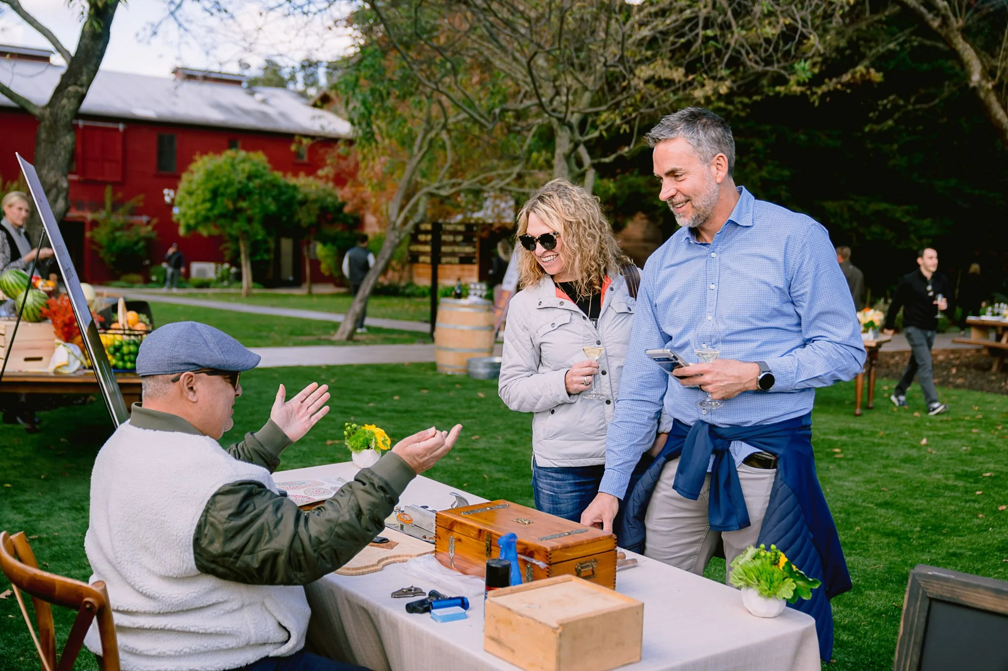 Two women and a man are smiling and chatting while standing near a table outdoors during fall. The table has a small wooden box, some tools, and a potted plant. There are people and a red barn in the background at what appears to be a farmers market 