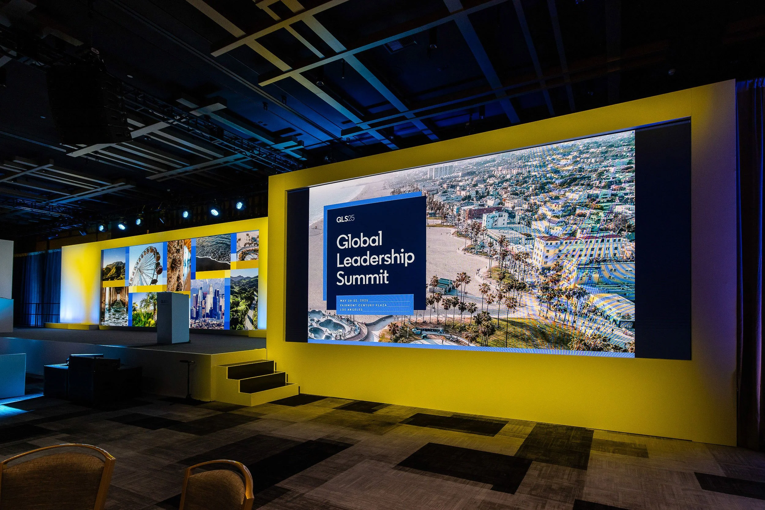 A large conference stage with a yellow border and multiple screens showing images of urban scenes, palm trees, and the Gulf of Mexico, at the Global Leadership Summit event, held in a spacious room with dark ceiling and blue lighting.
