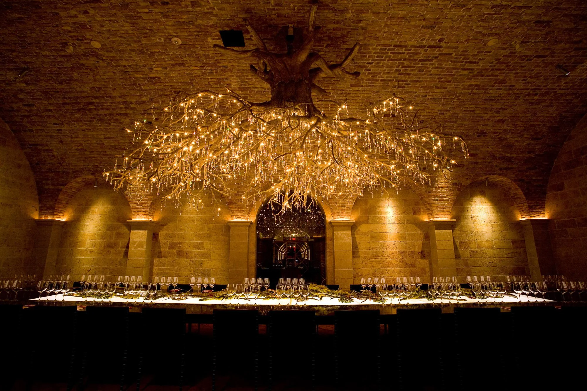 Elegant dining room with long table set with wine glasses and greenery, featuring a large chandelier made of tree branches with hanging crystals, against a brick arched ceiling.