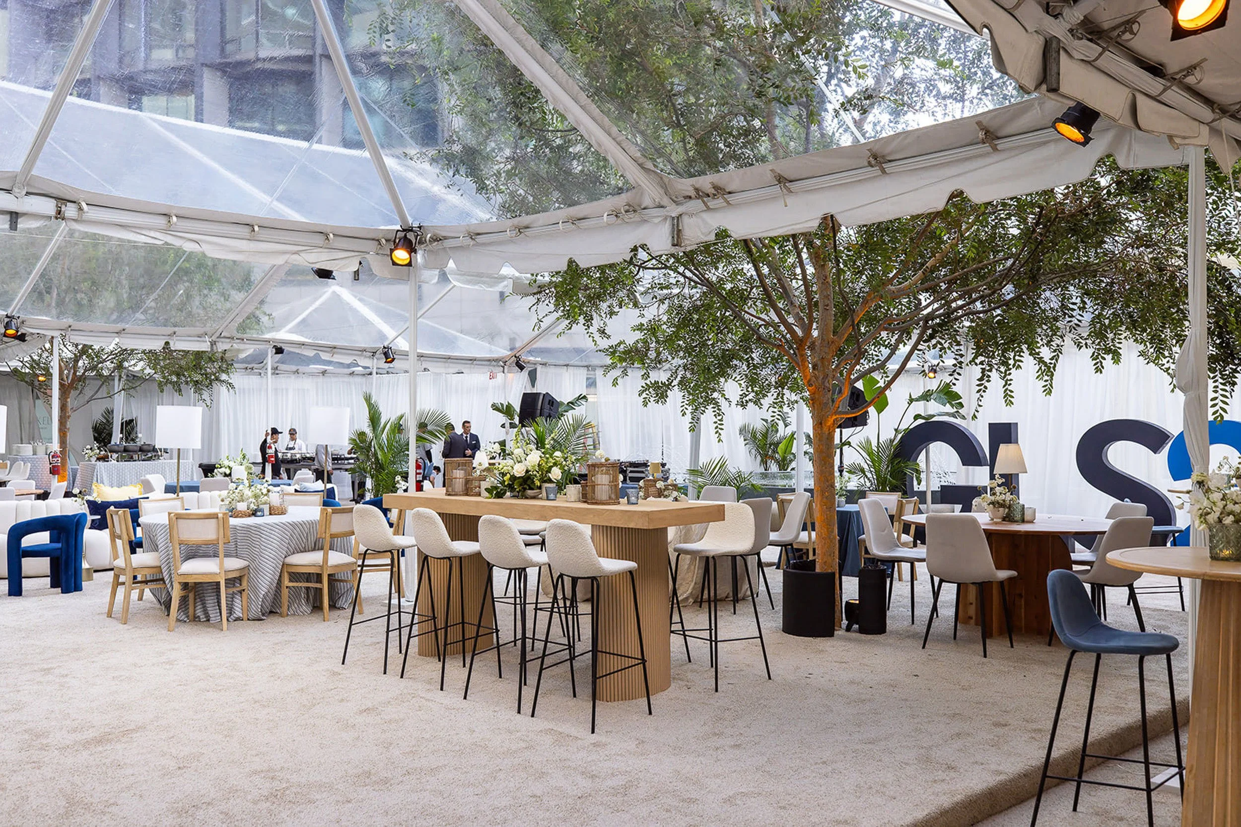 Indoor event space with tables and chairs, decorated with flowers, and greenery trees inside a transparent tent ceiling.