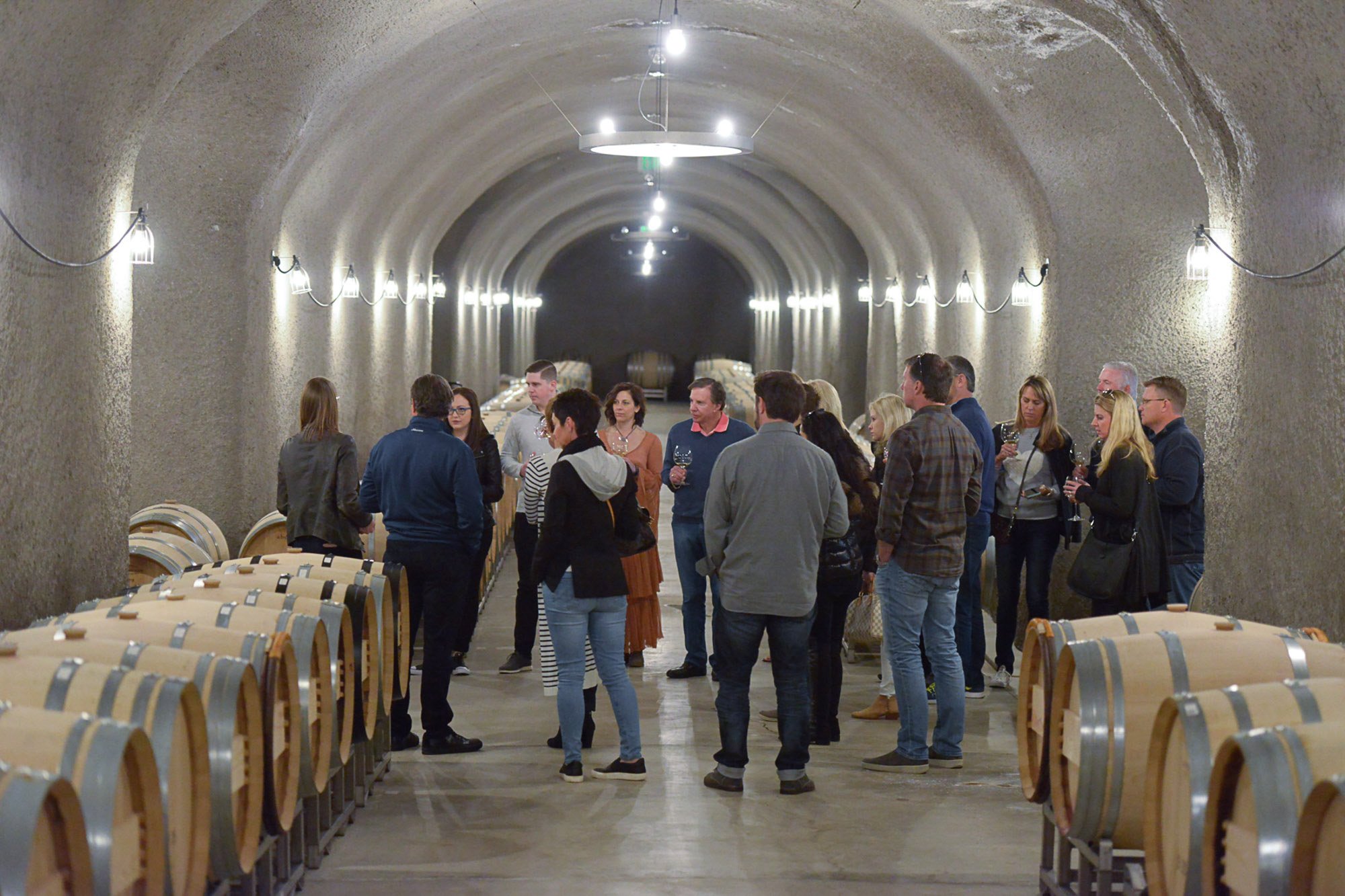 A group of people in a wine cellar, tasting wine from glasses, surrounded by wooden wine barrels.