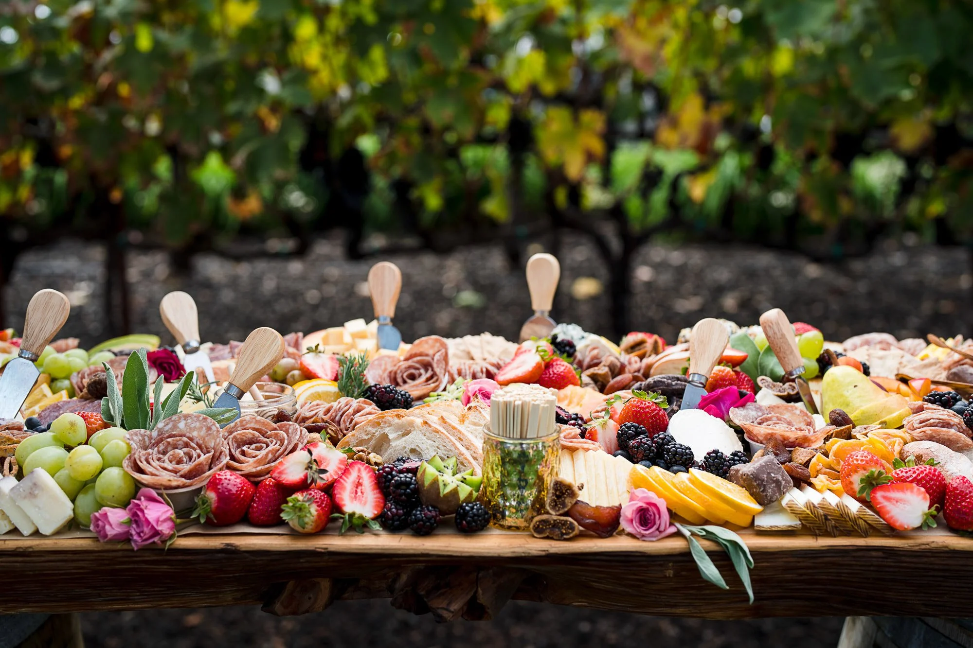A large wooden table filled with an assortment of cheeses, fruits like strawberries, grapes, and blackberries, sliced meats, and decorative flowers, set outdoors in a garden area with green plants in the background.