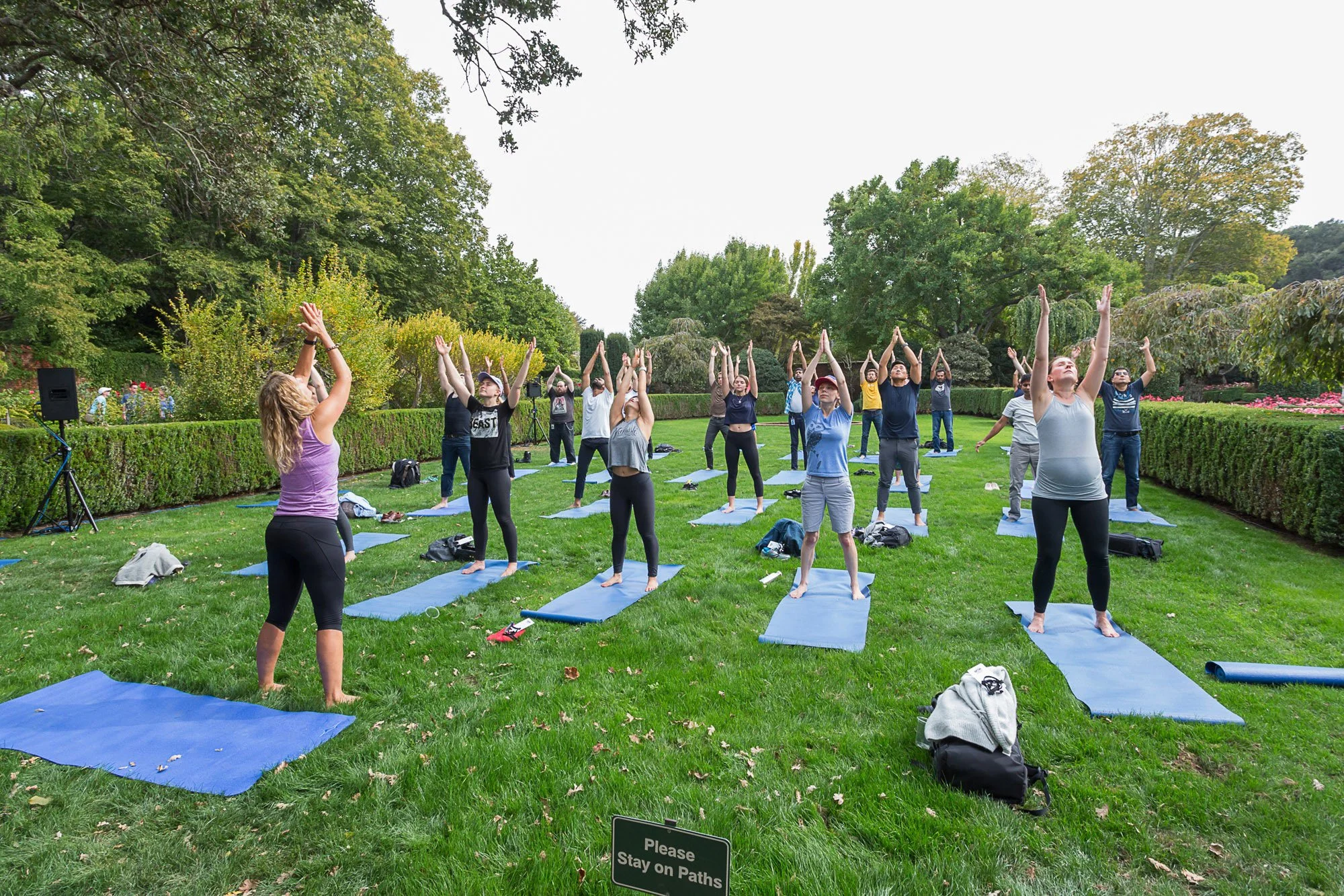 A group of people participating in a yoga class outdoors in a park, standing on yoga mats, with trees and bushes in the background.