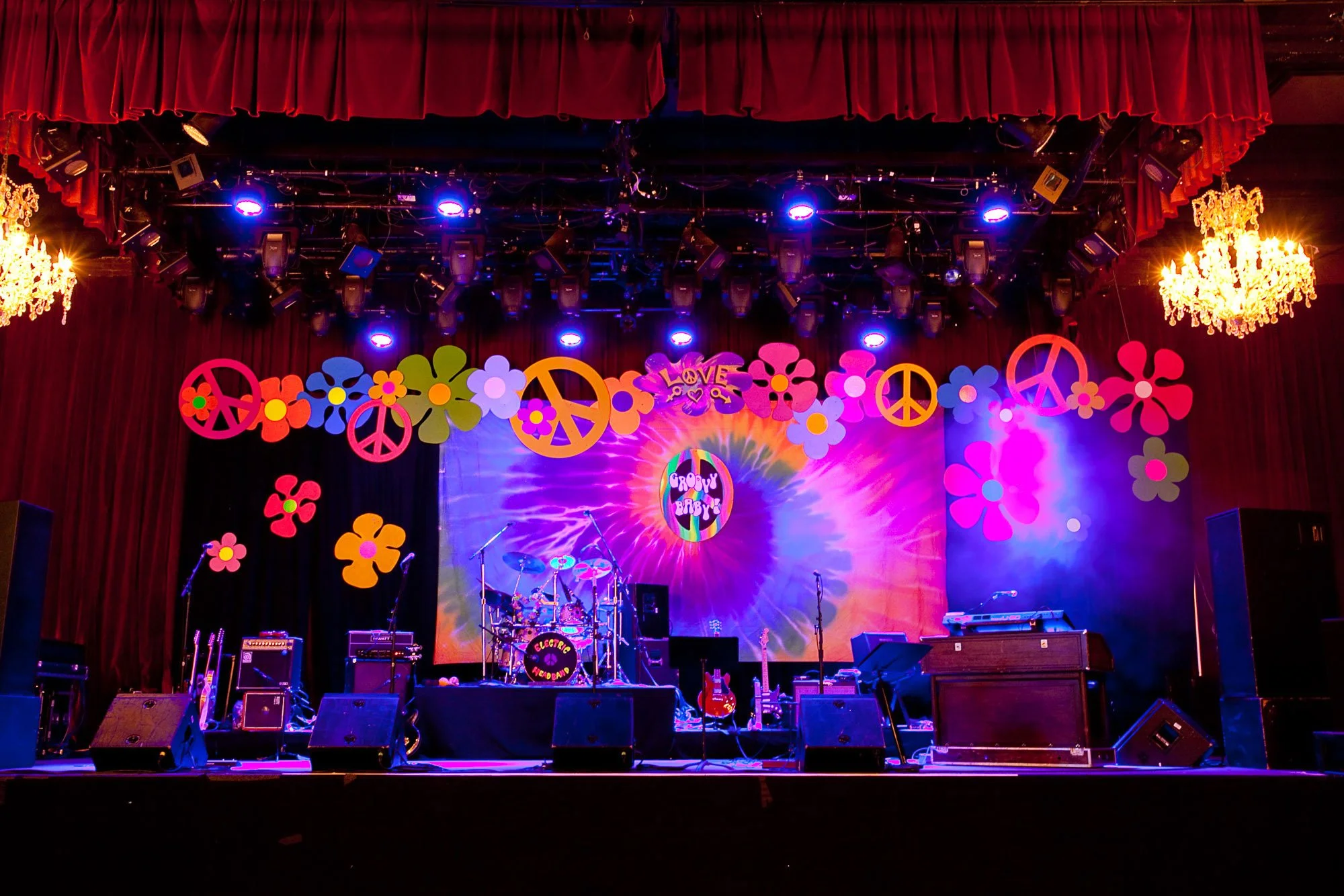 Stage set up for a music concert with colorful flower and peace sign decorations, purple and pink lighting, musical instruments, and equipment.