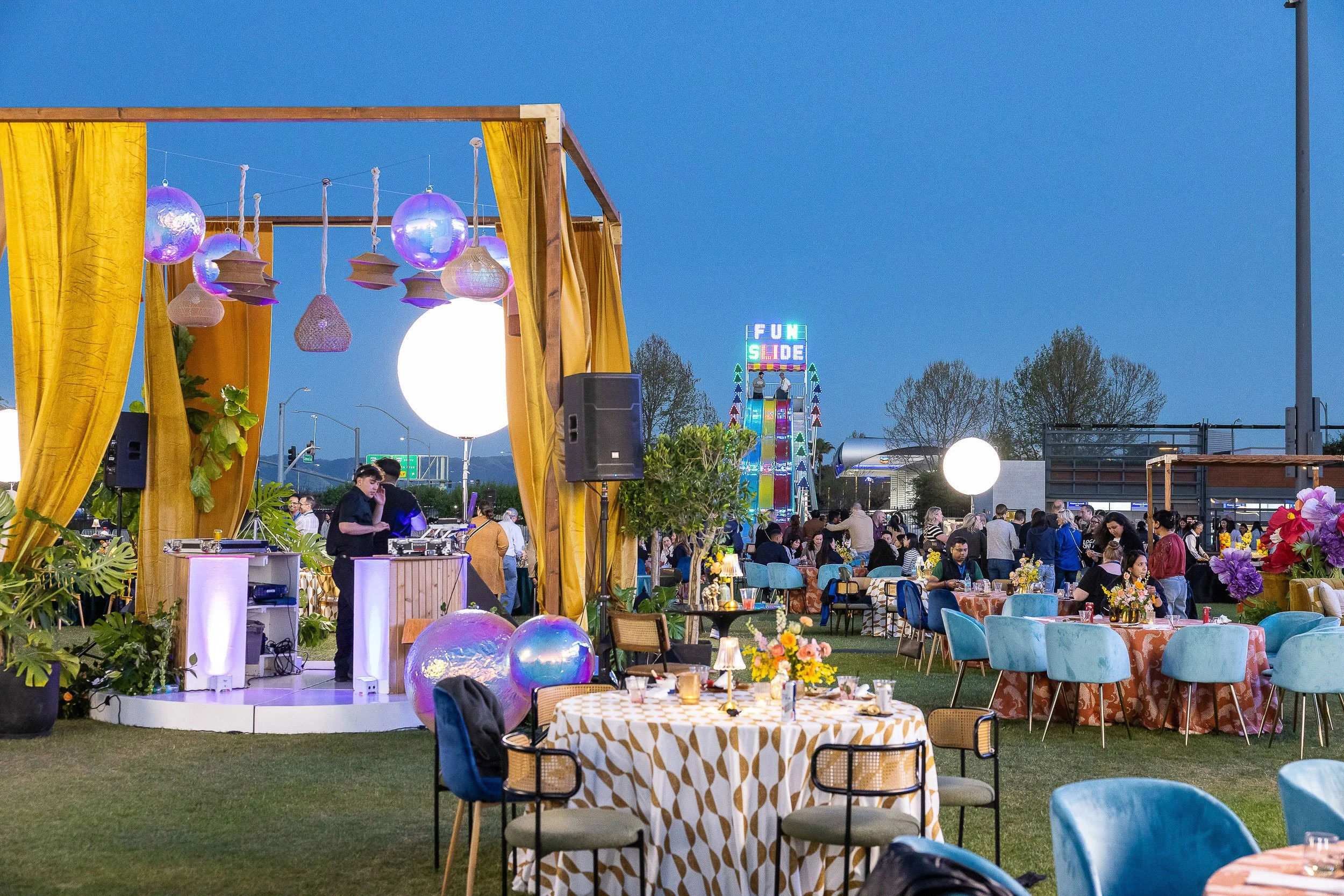 Outdoor event at dusk with colorful decorations, tables with checkered tablecloths, a variety of chairs, a carnival ride in the background, and a stage with hanging lanterns and lights.