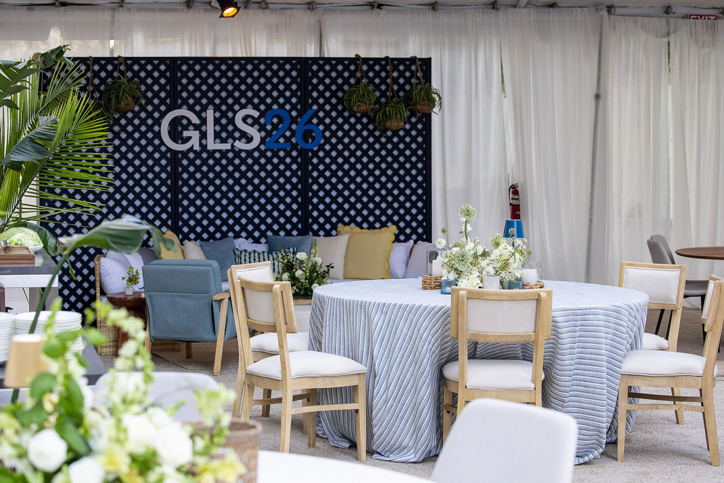 Indoor event space with a black lattice backdrop displaying 'GLS26', decorated with hanging plants. Round tables with floral centerpieces and a variety of chairs, surrounded by white curtains.