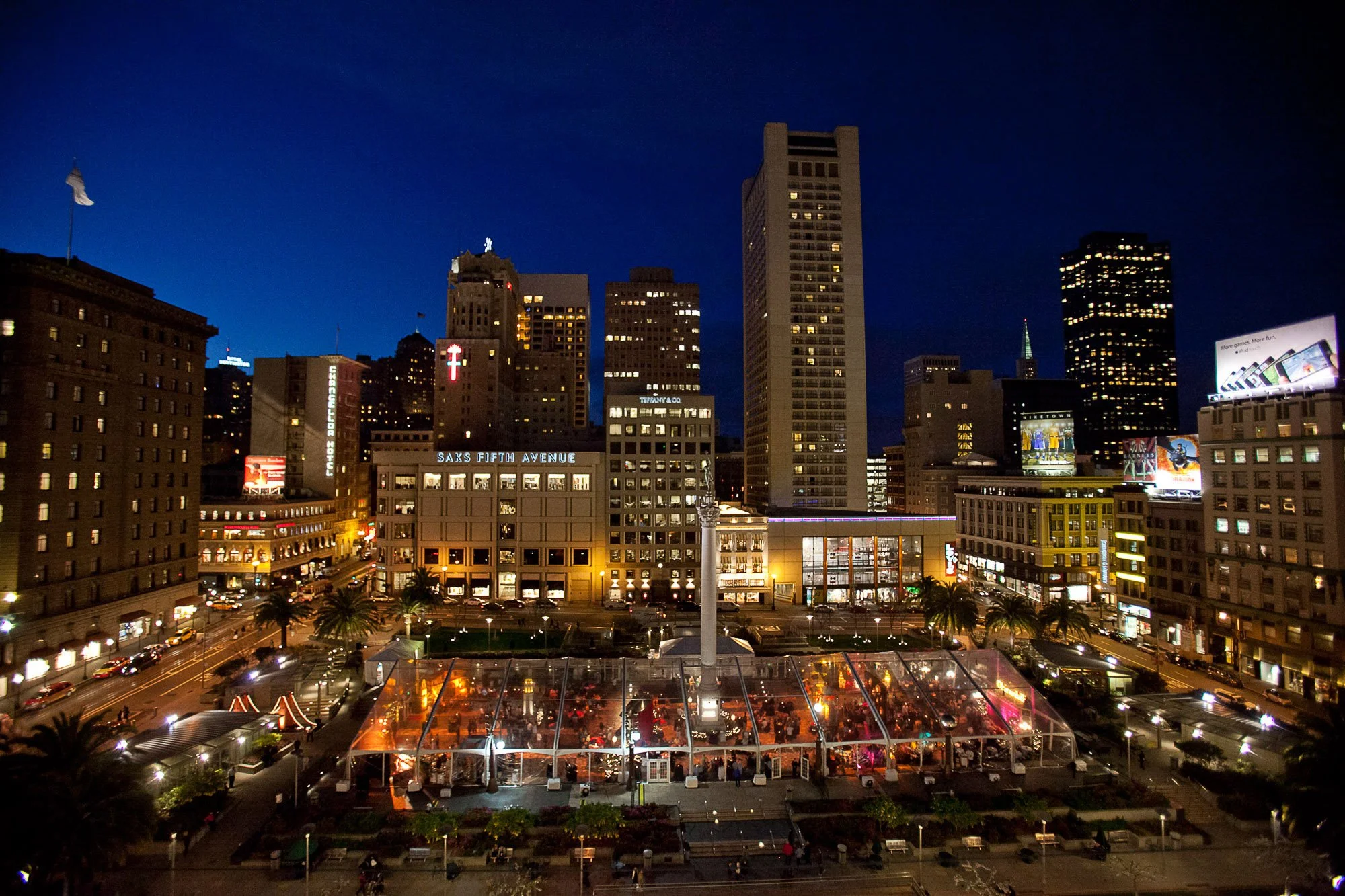 Night city skyline with tall buildings, illuminated windows, Billboards, and the San Francisco city hall in the foreground with a glass-roofed event space.