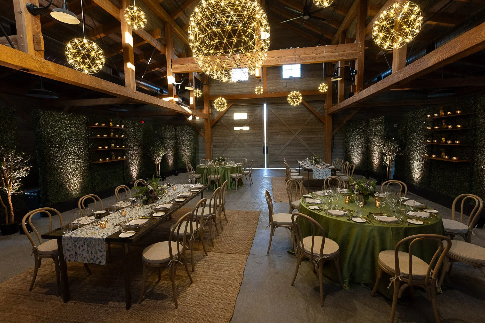 A decorated rustic indoor event space with hanging spherical string lights, round tables with green and patterned tablecloths, place settings, and floral centerpieces. The space has wooden beams and walls with ambient lighting.