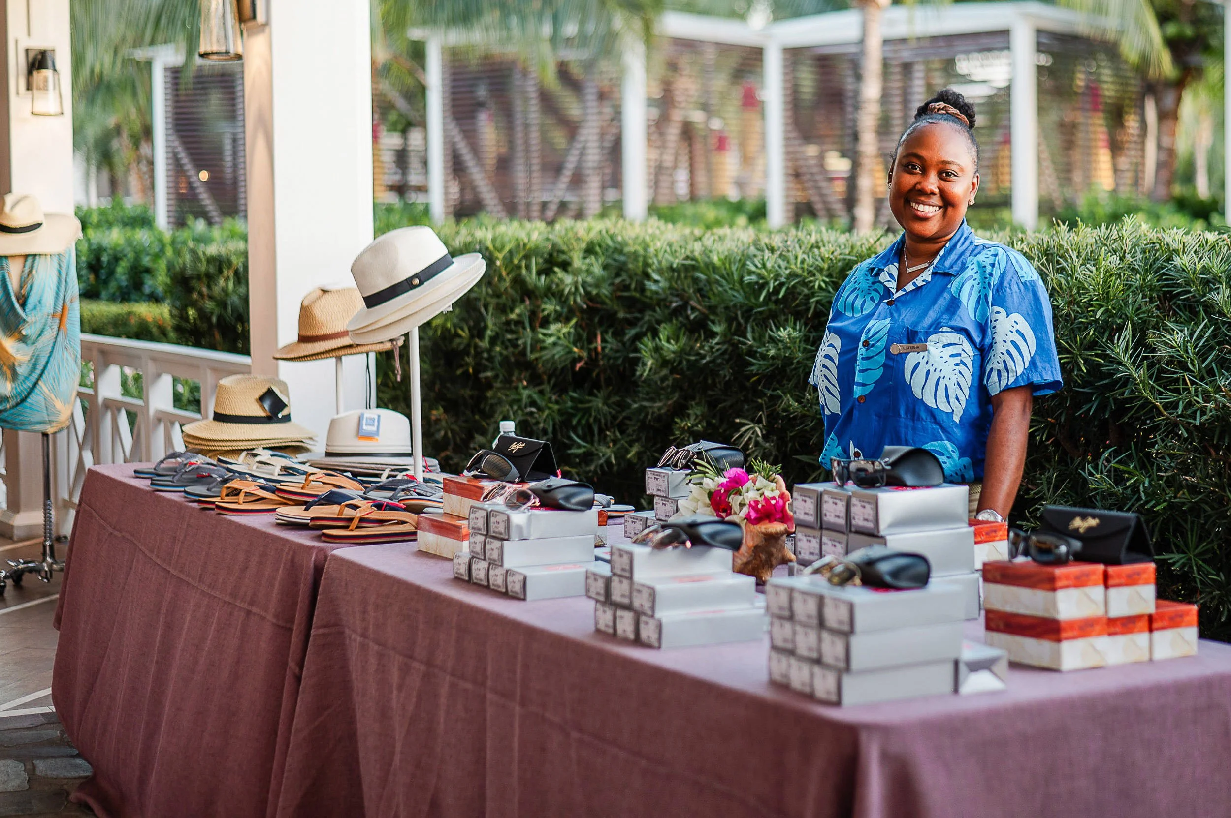A woman wearing a blue floral shirt stands behind a table displaying hats, sunglasses, and jewelry at an outdoor market with greenery in the background.