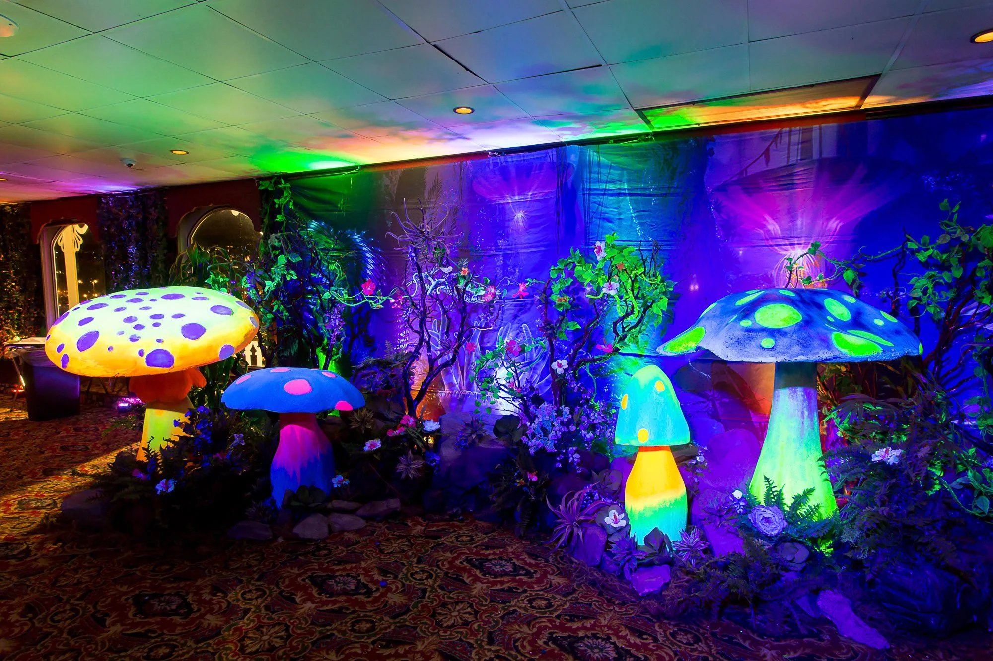 Colorful illuminated mushroom decorations with plants and flowers in the background on a carpeted floor.