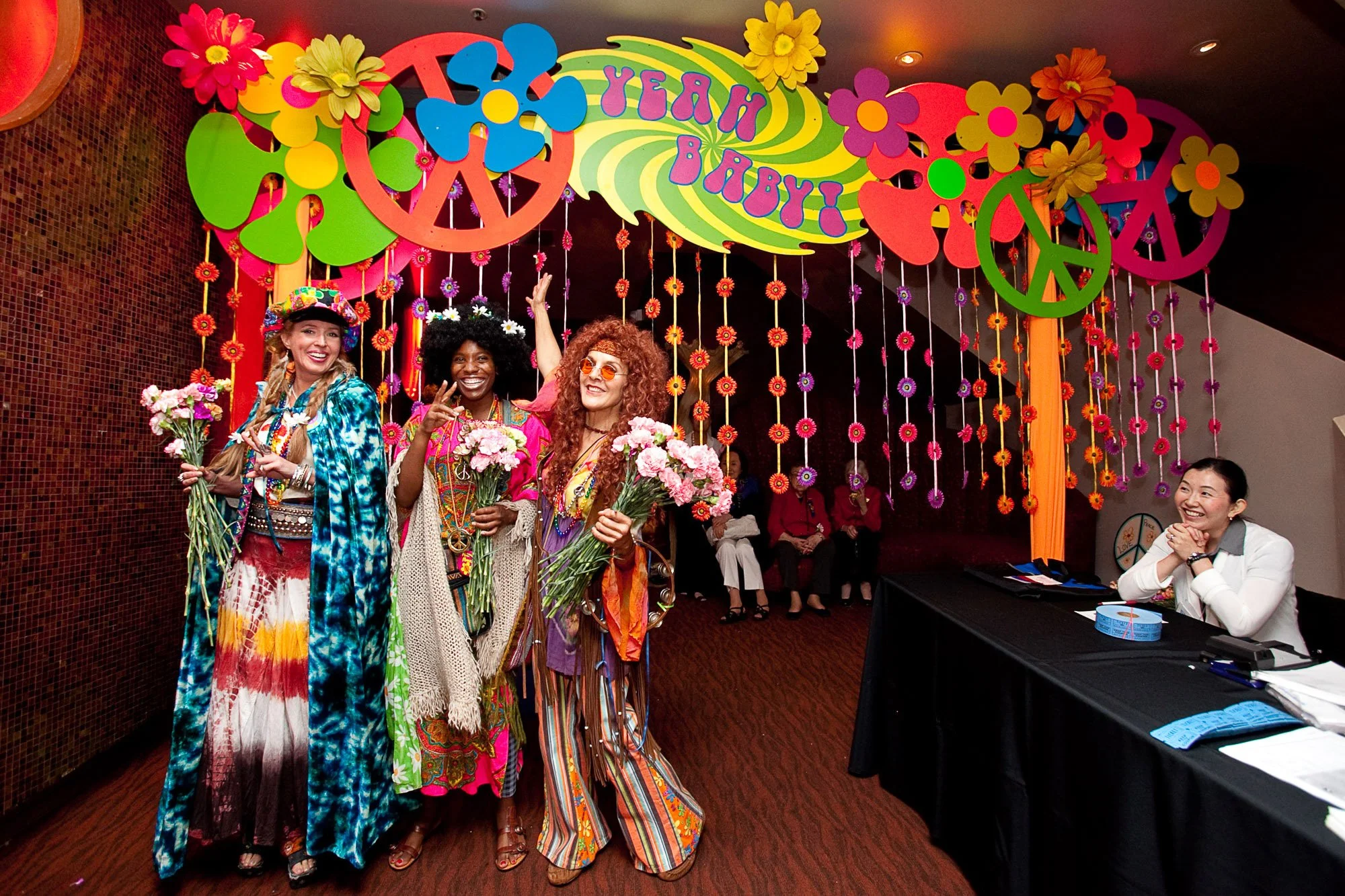 Three women dressed in colorful 1960s hippie-style outfits holding flowers, standing in front of a vibrant sign that reads '55 Year Baby!' with peace symbols, flowers, and psychedelic designs. Several people are seated in the background, and a woman 