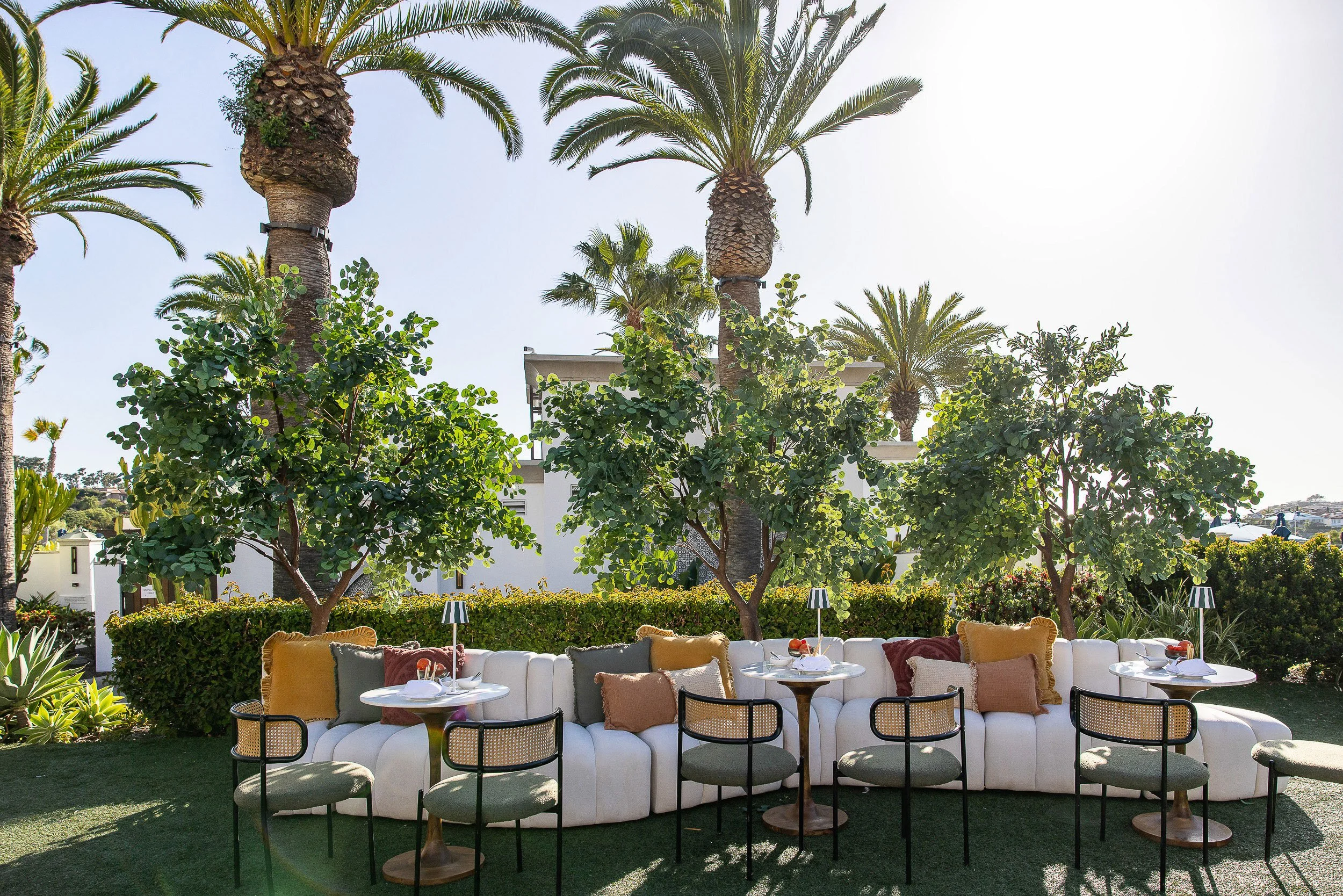 Outdoor seating area with a white sofa, yellow cushions, tables with place settings, and surrounding greenery including palm trees and hedges.