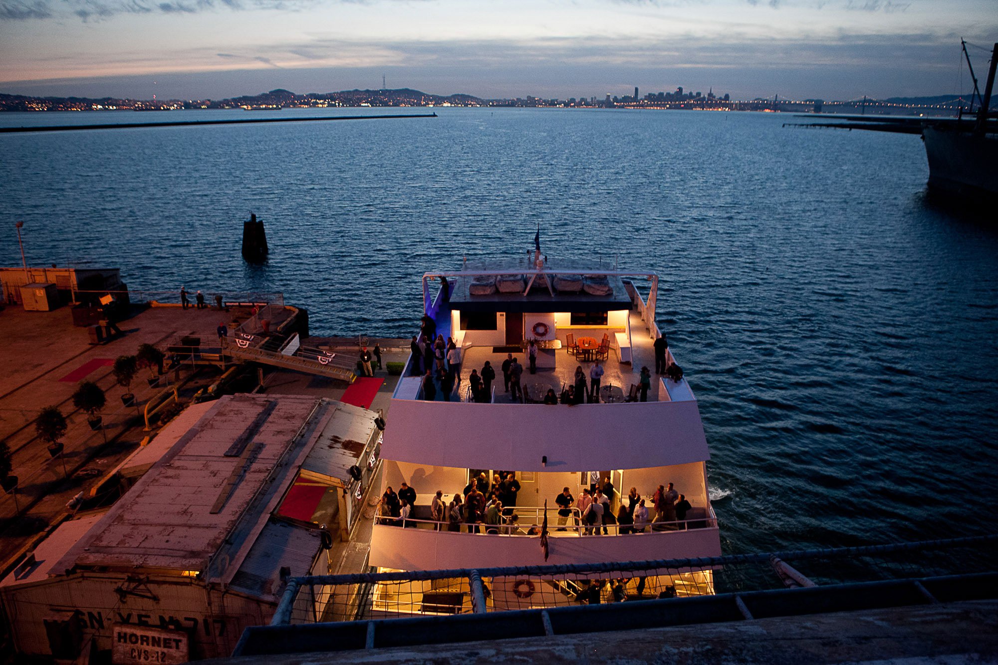 A large observation deck on a yacht with people socializing during sunset, with a city skyline and a bridge in the distance across a body of water.