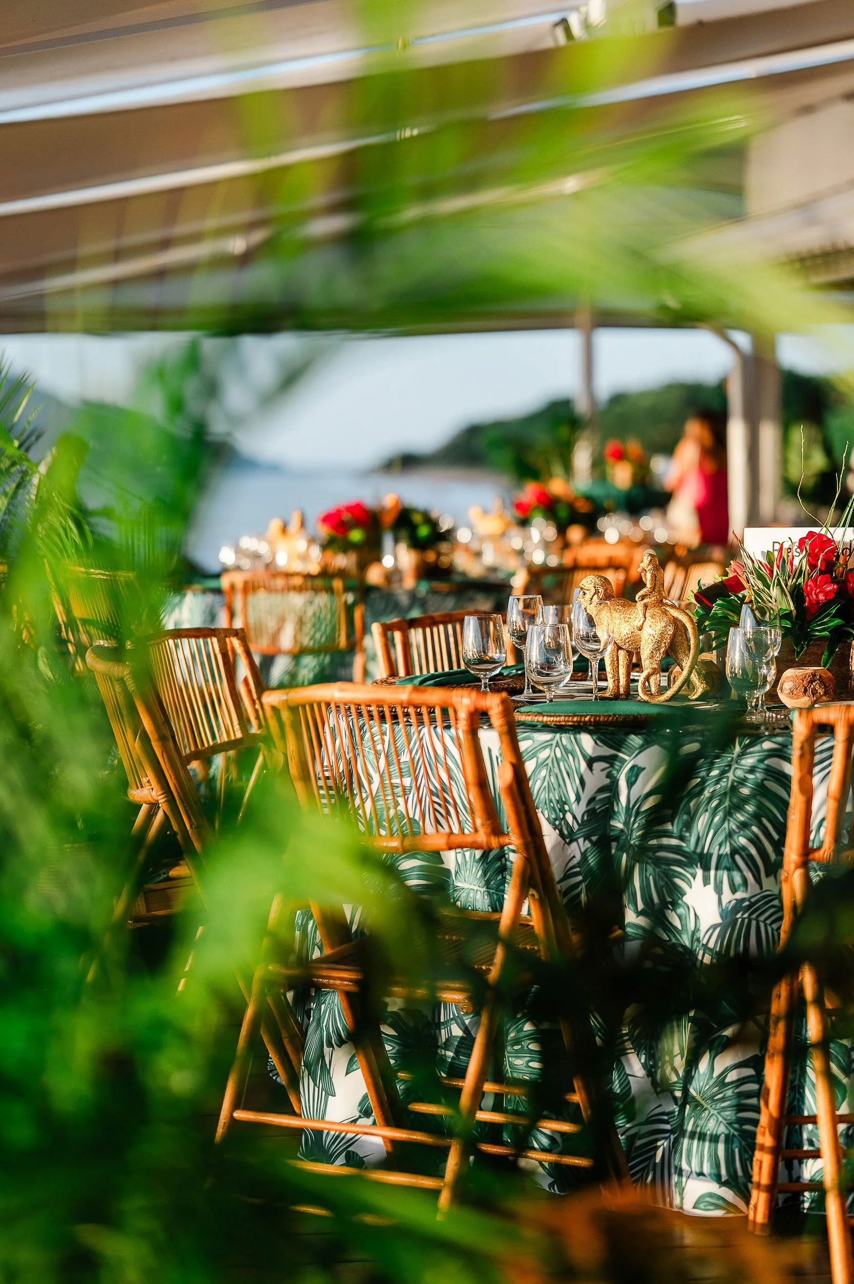 A tropical outdoor dining setup with a table decorated with a tablecloth featuring green and white leaf patterns, surrounded by wooden chairs. The table has flower arrangements with red flowers and decorative elephant statues. There are wine glasses 