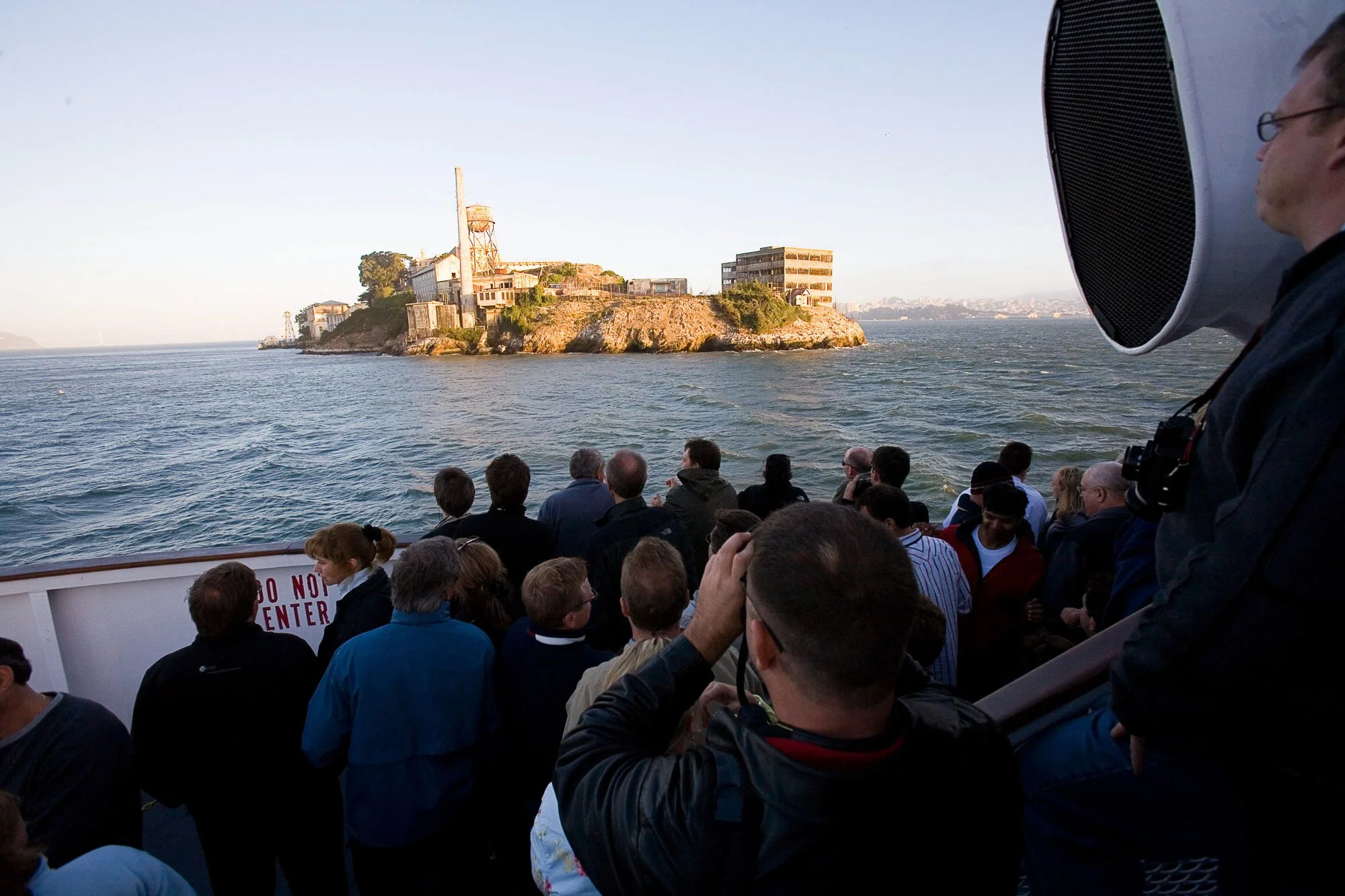 A large group of people are on a boat, looking at Alcatraz Island in the distance. The island features buildings and a water tower, with the bay surrounding it and a clear sky above.