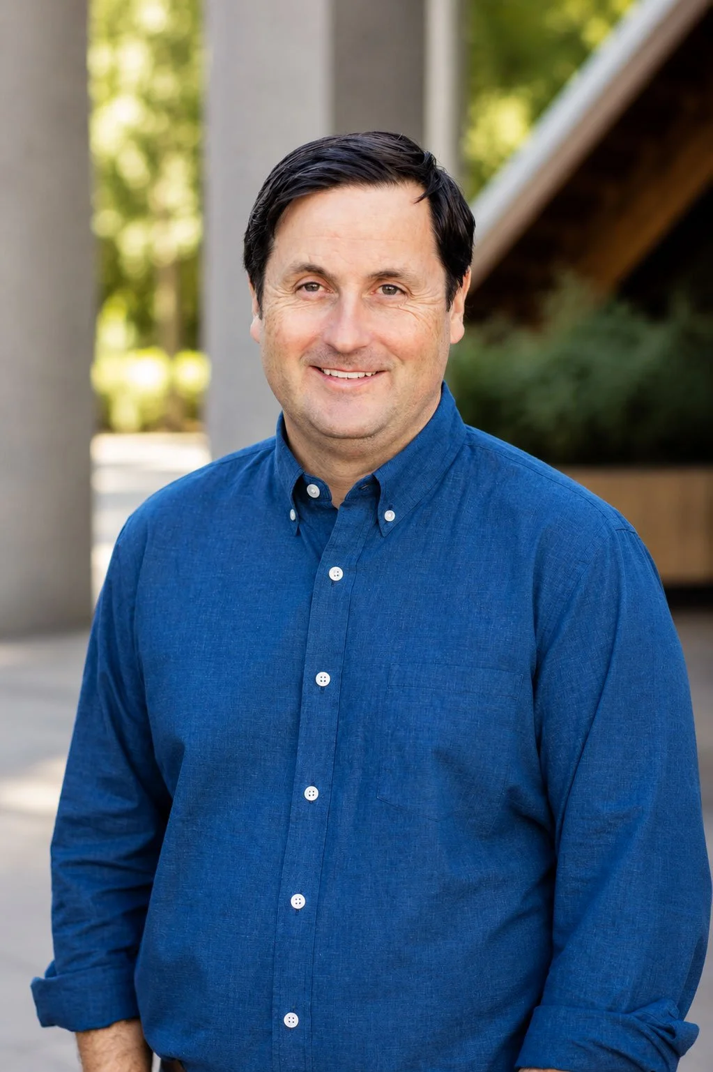 A man with dark hair wearing a blue button-up shirt, smiling outdoors with trees and a structure in the background.