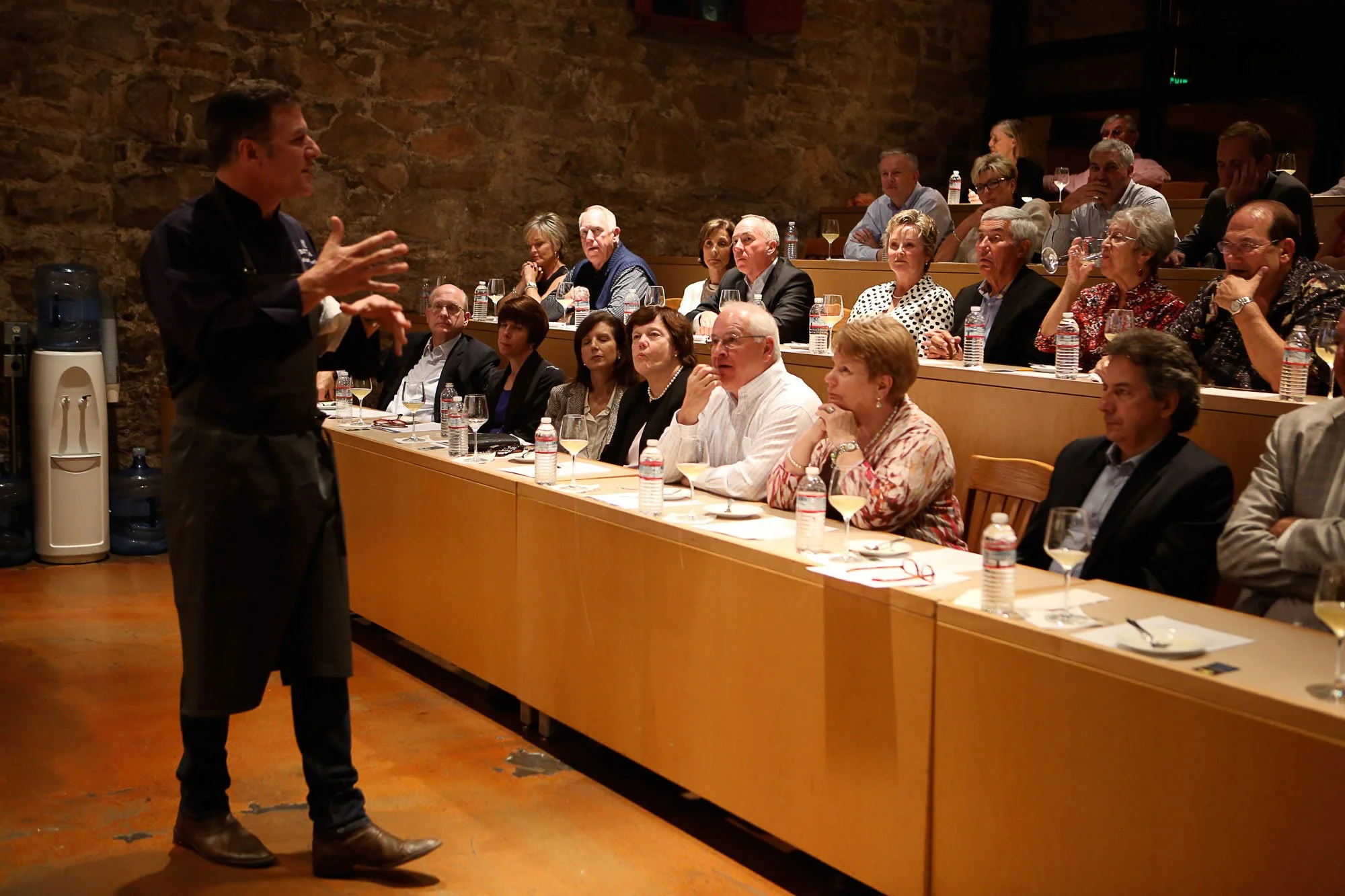 A man giving a presentation or speech to an audience of mostly middle-aged and older adults seated at tables. The setting appears to be a conference or seminar room with a stone wall.