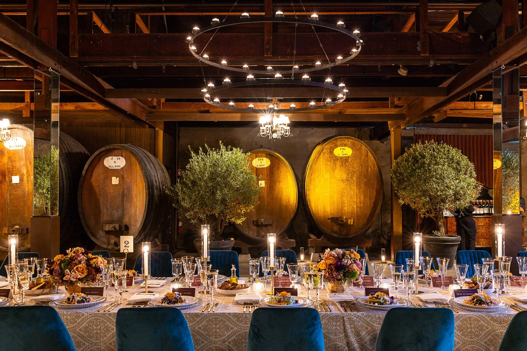 Elegant dining table set for a formal event with flower centerpieces, candles, and glassware inside a rustic venue with large wine barrels and wooden beams.